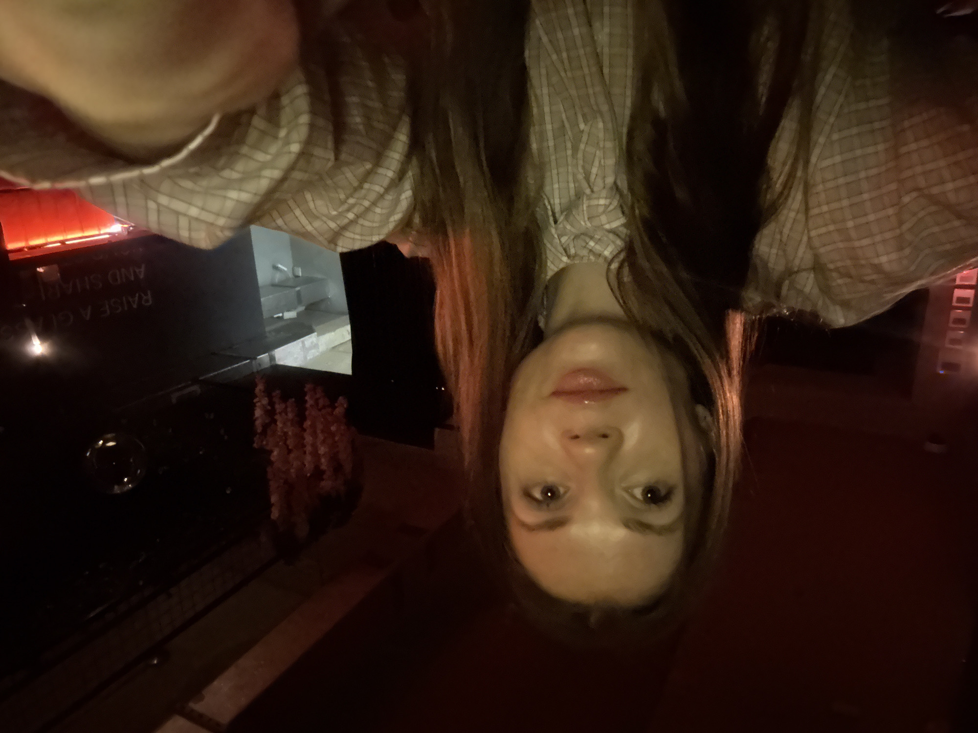 A female stands upside down in front of a bar in Batumi, Georgia. The bar has a sign that reads "Raise a glass and share." There are flowers hanging and tables in the background. The woman has brown hair, fair skin, and is wearing a plaid shirt.