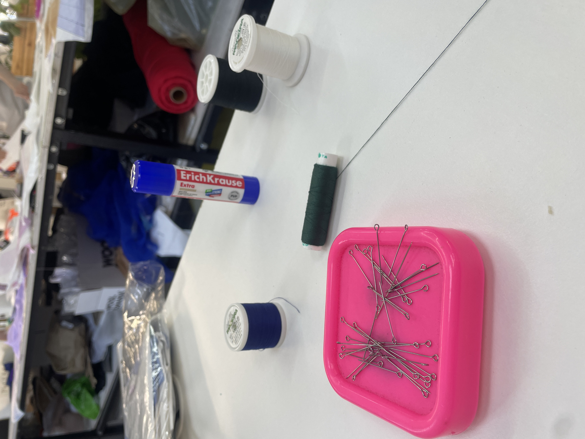 The image shows a close-up of a white table in what appears to be a workshop or craft area. On the table, there are several spools of thread: one blue, one black, one white, and one dark green. A bright pink tray filled with sewing pins is in the foreground. To the left of the spools, there is a blue and white tube, possibly glue or a marker, with the text "ErichKrause Extra" visible. In the background, partially obscured by the table and other objects, is a shelf unit containing various items, including fabric rolls, paper products, and what looks like plastic sheeting. There are no people directly visible in the frame, but the presence of tools and materials suggests ongoing work. The lighting suggests an indoor environment, possibly with overhead lighting. The scene is clean and organized, with the objects neatly arranged on the table. The overall impression is one of a practical workspace. The location of Pargolovo, Russia, is not visually ascertainable from the image itself; this information is provided externally.