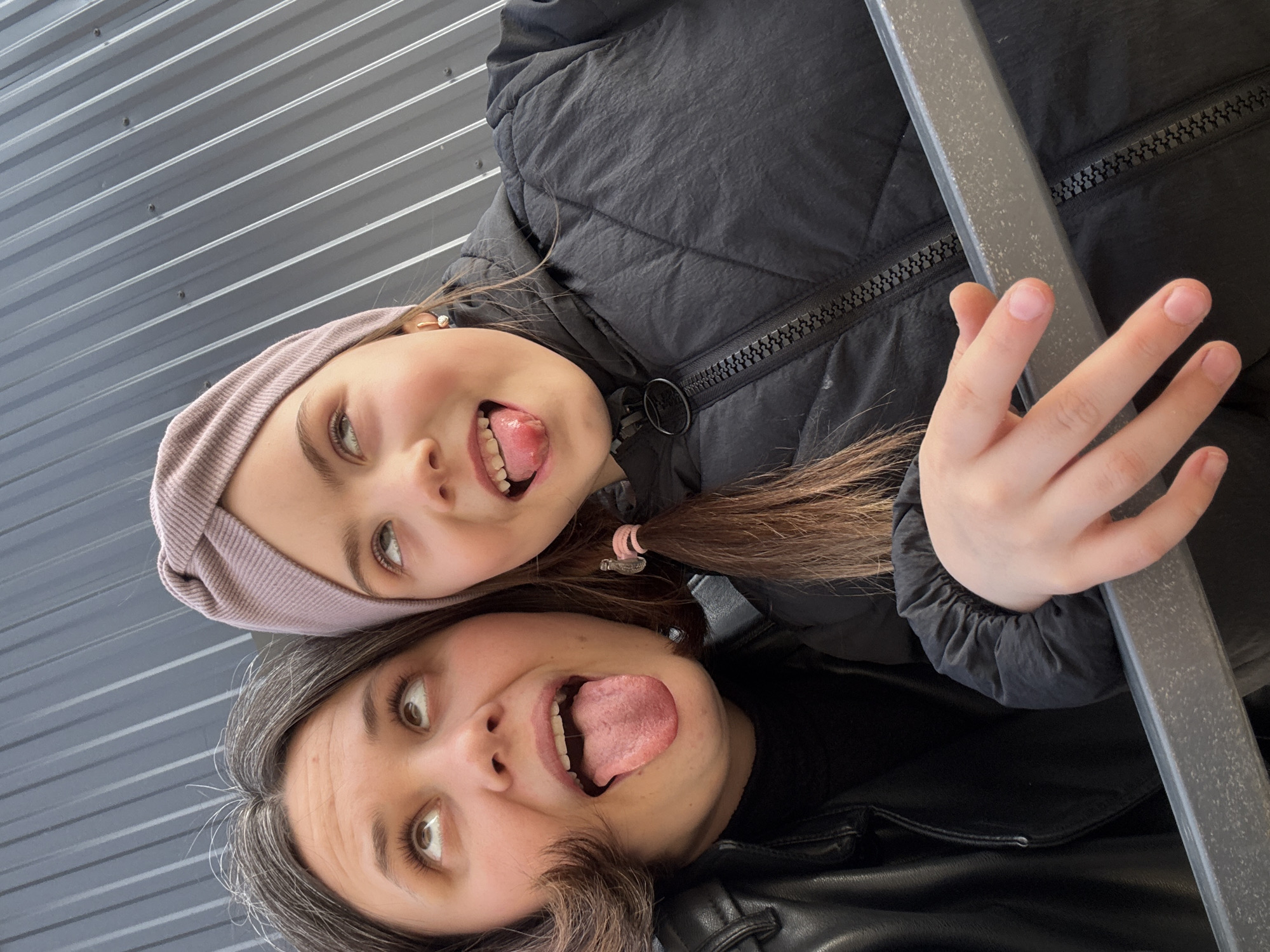 Two individuals, a child and an adult, are posing for a photograph. The child is positioned above the adult. Both have their tongues extended in a playful manner. The child wears a lilac-colored head covering and a black puffy coat. The adult wears a black leather jacket. The photo appears to be taken in an angled or sideways manner. A gray metal beam or pole is partially visible on the right side of the image. The background consists of horizontally lined gray panels. The photo's location is identified as the city of Chaikovsky, Russia.