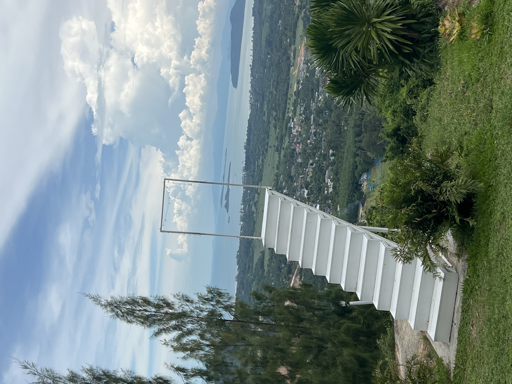 The image depicts an outdoor scene, likely a viewpoint, overlooking a landscape. In the foreground, a striking white staircase with evenly spaced steps dominates the right side of the frame, ascending diagonally. The staircase appears to be an architectural element, possibly for viewing or accessing a higher point. A thin metal structure, possibly a frame or support, is visible at the top of the stairs, adorned with what looks like a string of lights.
Beyond the staircase, the scene opens up to a panoramic view. A vast body of water, likely a lake or the sea, stretches across the middle ground, with islands or peninsulas dotting its surface. The water is a muted blue-gray, reflecting the sky. The distant shoreline is lined with lush green vegetation and a scattering of buildings, suggesting a populated area or a town nestled amidst the trees. The landscape recedes into the distance, where hazy mountains can be seen on the horizon.
The sky is filled with dramatic, billowing white clouds against a bright blue backdrop, indicative of a sunny day with some cloud cover. The lighting suggests it is daytime, likely late morning or afternoon. The overall impression is one of a scenic overlook, offering breathtaking views of nature and human settlement. The composition, with the stark white staircase juxtaposed against the natural landscape and sky, creates a visually engaging contrast. The image is oriented vertically, with the top of the scene appearing on the left. The location context provided is Baan Thurian, Thailand. There is no visible text in the image.