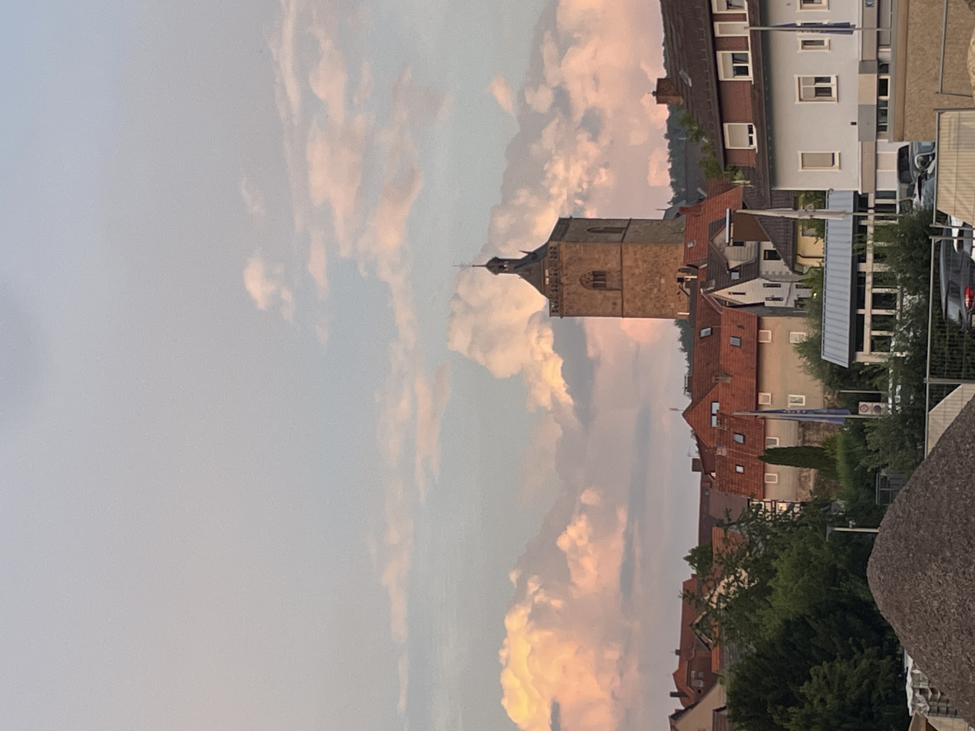 The image depicts a townscape with a prominent church tower in Neukirchen, Germany. The sky is filled with pink and orange clouds, suggesting either sunrise or sunset. Several buildings with red-tiled roofs are visible in the foreground and middle ground. The church tower is made of stone and has a cross at its apex. In the lower part of the image, there is a pile of gravel and construction materials, indicating ongoing work or development in the area. Some trees and greenery are interspersed among the buildings.