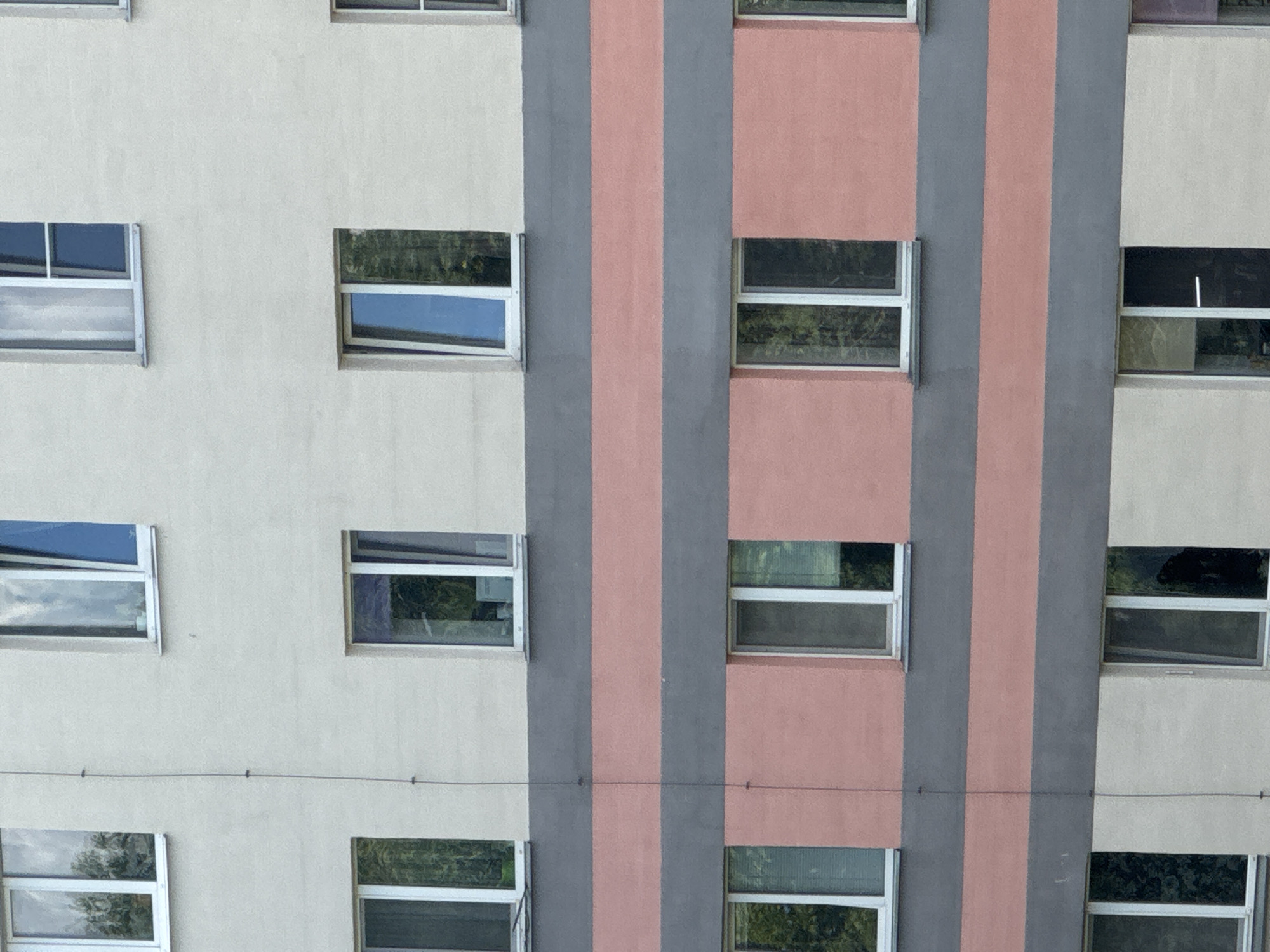 The image shows the exterior of a multi-story building with a repetitive pattern of windows and horizontal bands of color. The building's facade is divided into sections of cream, gray, and pink. There are multiple windows visible, all with white frames. Some windows are open, while others appear to be closed. Reflections in the windows suggest the presence of trees and sky. The overall impression is of an apartment building or similar structure in an urban or suburban environment. The location is specified as Яромінскі сельскі Савет, Belarus.