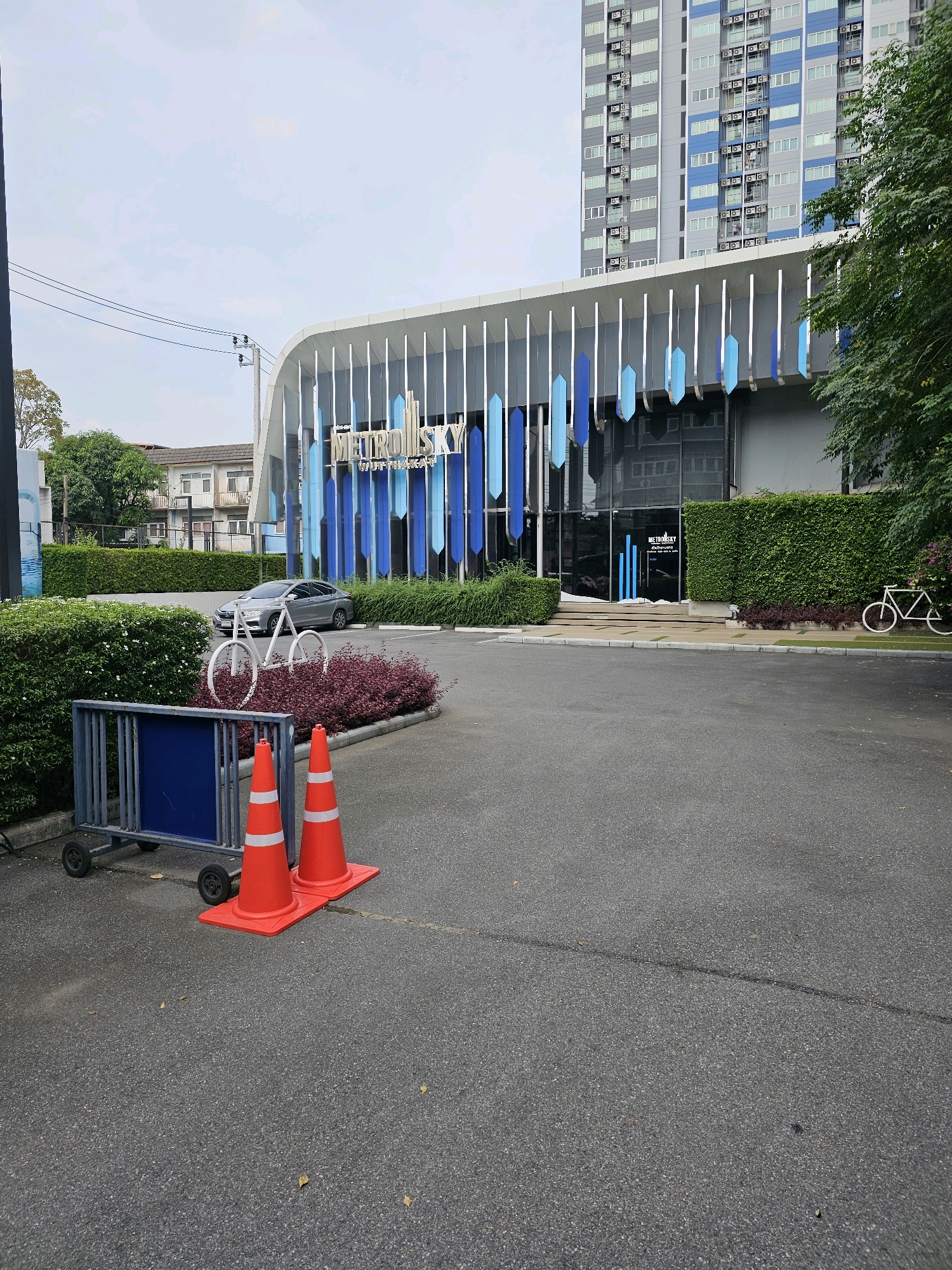 In the city of Bangkok, Thailand, the image captures a frontal view of the METRO SKY WUTTHIKAT condominium building. The condominium features a modern architectural design with a curved white facade and vertical blue accents. In front of the building, a neatly manicured hedge lines the property. A silver sedan is parked on the asphalt driveway, and a white metal bicycle sculpture is placed near the hedge. Two orange traffic cones with white reflective stripes are positioned on the asphalt in the foreground, next to a metal barricade with wheels. The sky is overcast, and trees are visible in the background.
