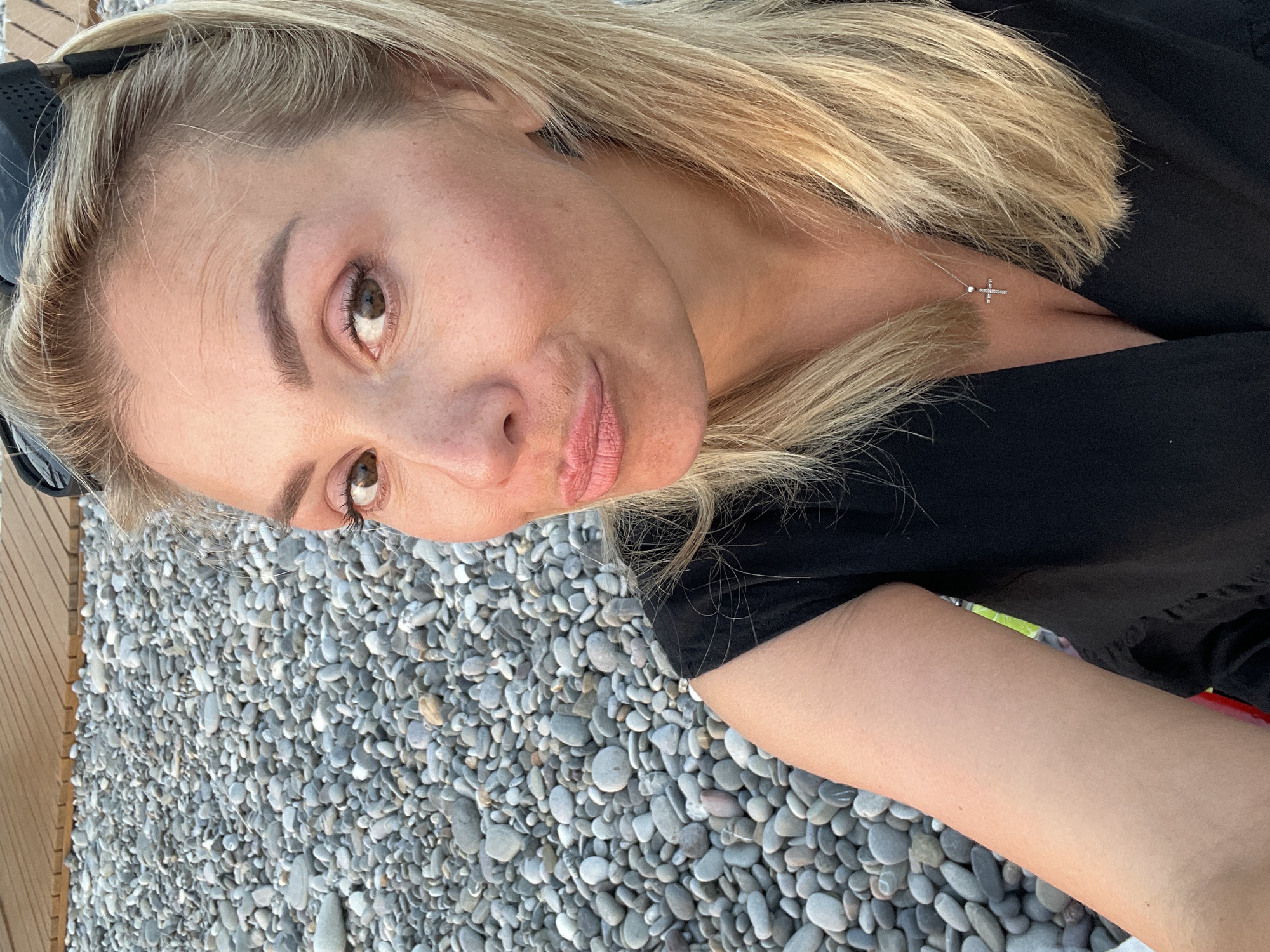 A woman with blonde hair and brown eyes is taking a selfie on a pebble beach. She is wearing a black top and a silver cross necklace. The background is a mix of pebbles and a wooden deck. She is making a duck face.