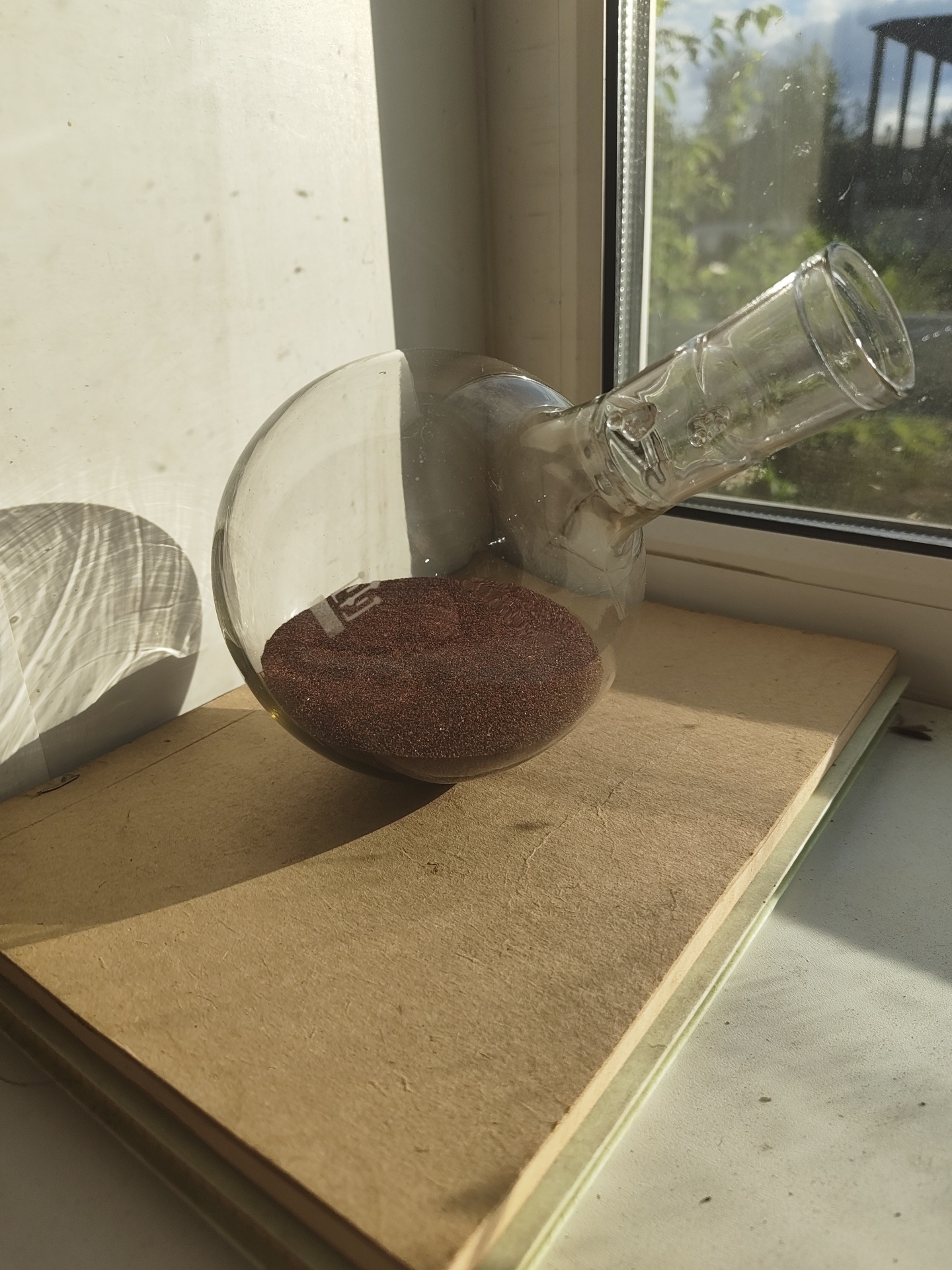 A round-bottomed glass flask filled with dark reddish-brown granular material is positioned on top of a stack of books. The flask's neck extends upwards and to the right, angled towards a window. Sunlight casts a distinct shadow of the flask's curved body onto a white wall to the left of the frame. The scene is illuminated by natural light filtering through the window, which shows greenery and a building exterior in the background. The books appear to be old, with textured brown covers. The overall environment suggests an indoor setting, possibly a windowsill or desk, in Metallostroy, Russia. There are no people or animals present.