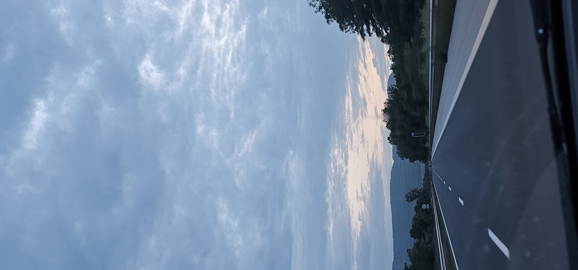 The image depicts a serene, late afternoon or early evening scene captured from a moving vehicle on a road. A wide, dark asphalt road with distinct white lane markings extends from the bottom of the frame and curves slightly into the distance towards the upper right. Lush green trees and vegetation line the right side of the road, forming a dense border. Beyond the trees, in the mid-ground, a range of dark, undulating hills or mountains is visible against the horizon.
The sky dominates the left and upper portions of the image, filled with a soft blue hue and scattered, light gray and white clouds. Some clouds are wispy and streaky, while others are more defined and billowy. A faint, soft pinkish-orange glow is discernible along the horizon line, nestled between the distant mountains and the cloud-filled sky, suggesting either an approaching sunset or very early sunrise. Part of the car's interior or window frame is visible along the far right edge of the photograph. The overall atmosphere conveys a sense of peaceful travel. No discernible text is clearly visible, though a very faint, small object with "01" might be present on the left side of the road, partially obscured by trees.