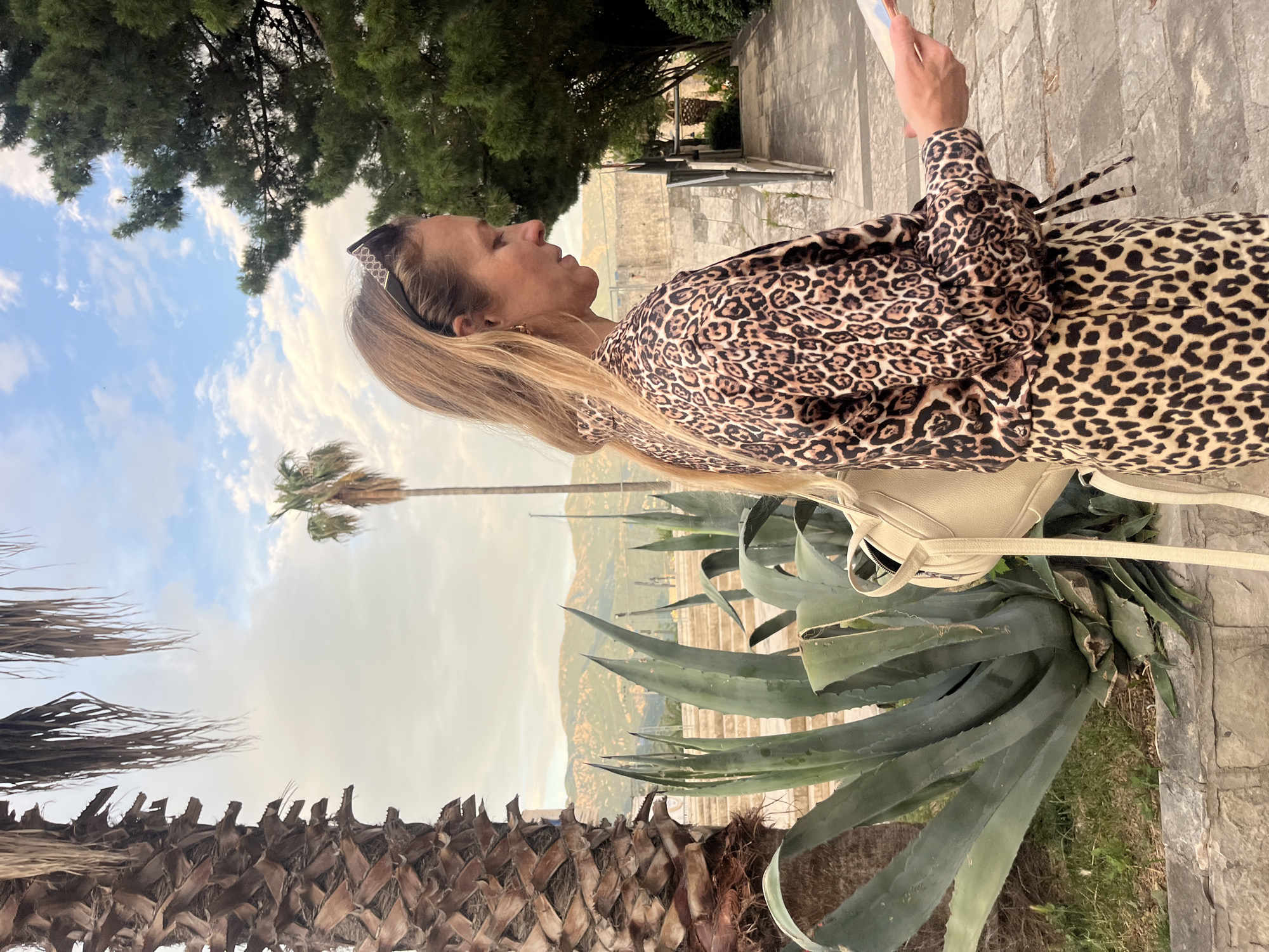 A side-view shot depicts a woman in a leopard-print outfit standing outdoors, likely in Budva, Montenegro. The background features agave plants, palm trees, a mountain range, and a cloudy sky. A cream-colored purse hangs from her shoulder. She holds a phone or similar device in her hand. The surroundings suggest an urban or garden setting with stone structures visible.
