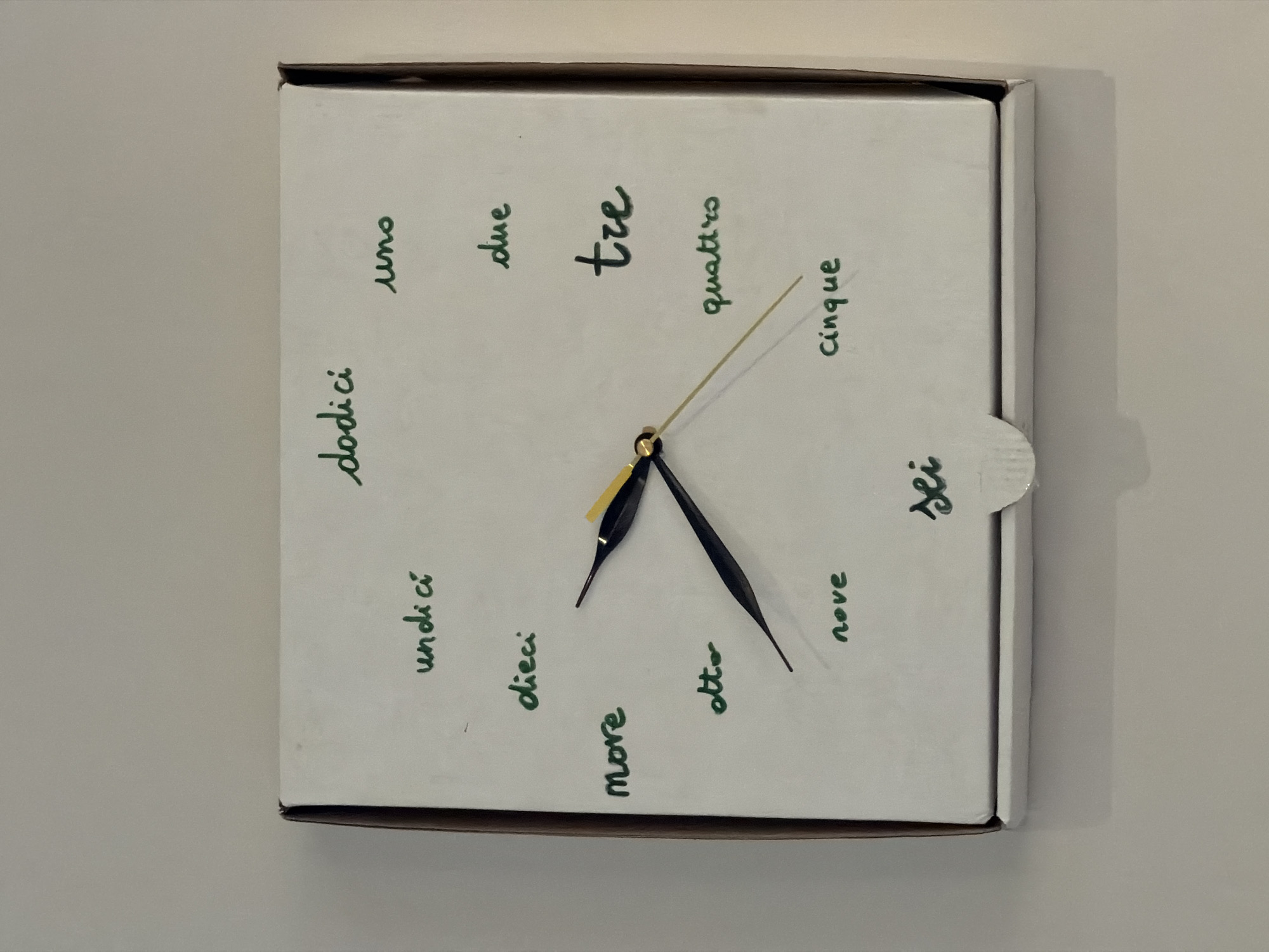 A top-down shot shows a white square clock hanging on a light-colored wall. The clock face is white with Italian words printed in green script around the perimeter to indicate the hours, starting with "dodici" at the top-left and going clockwise. The words and their approximate corresponding hours are: dodici (12), undici (11), dieci (10), nove (9), otto (8), sette (7), sei (6), cinque (5), quattro (4), tre (3), due (2), uno (1). The clock hands are black, with the hour hand pointing approximately to "dieci" (10) and the minute hand pointing near "otto" (8). The second hand is gold. The clock appears to be constructed from a white box. The clock is not located in Izhevsk, Russia.