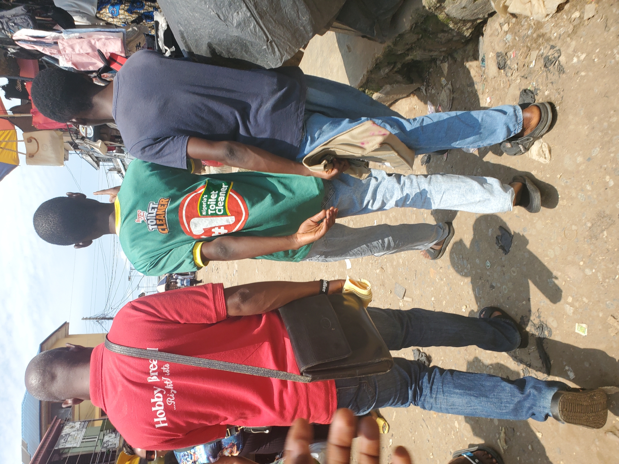 Three individuals are standing in what appears to be an outdoor market or street in Shangisha, Nigeria. The scene is characterized by a dirt or gravel ground with scattered debris. The lighting suggests it is daytime, likely sunny, with strong shadows cast by the people and other unseen objects.
The person on the far left is wearing a navy blue t-shirt and blue jeans. They are looking away from the camera. Next to them, in the center, is a person wearing a green t-shirt with yellow trim on the collar. This person is also looking away and has their arm outstretched, possibly gesturing. The green t-shirt has visible text and a graphic: "hypo TOILET CLEANER" in white text, with a red and white circular logo to the right that contains what looks like a toilet brush. The person on the right is wearing a red t-shirt with white text that reads "Hobby Breed... Right choice". This individual is also looking away from the camera. They have a black shoulder bag with a strap across their chest, and what appears to be a wallet or folder tucked into their jeans. All three individuals are wearing casual clothing and appear to be standing.
The background shows a cluttered environment with various items that might be merchandise or discarded materials, including fabrics and what looks like a large dark tarp or bag. There are also some buildings visible in the distance, and electrical wires crisscrossing overhead, indicative of an urban or semi-urban setting. The overall impression is a candid snapshot of daily life in a busy Nigerian marketplace.