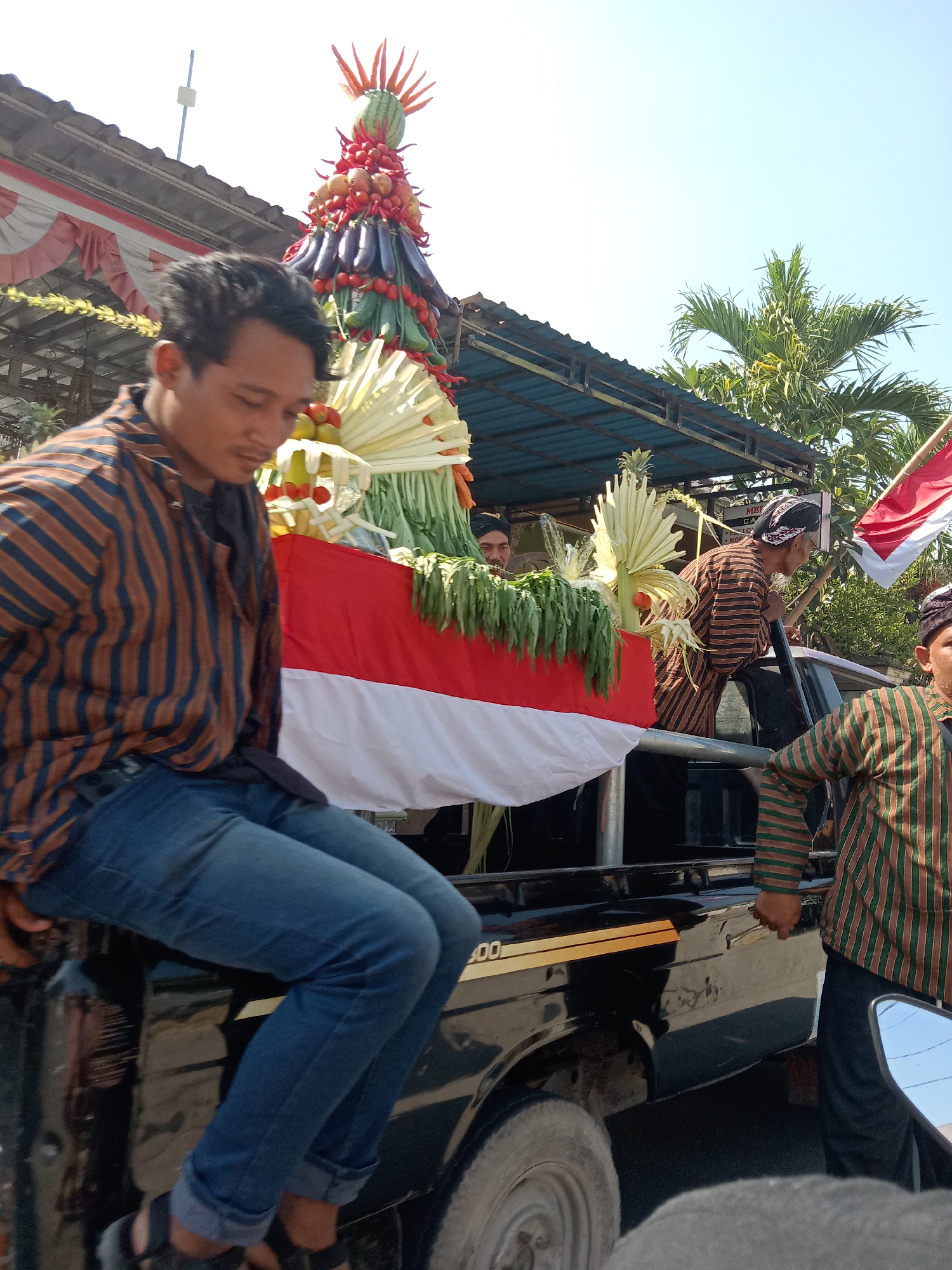 In the city of Alasgung, Indonesia, a procession is underway, with a truck serving as a moving platform for a large, conical arrangement of fruits and vegetables. The centerpiece of this arrangement appears to be a watermelon, crowned with chili peppers and adorned with eggplants and tomatoes. Beneath the produce, a red and white banner, likely the Indonesian flag, is draped.

On the truck, several individuals are present. In the foreground, a man wearing a striped shirt and blue jeans is seated on the edge of the truck bed, looking down. To his right, another man in a similar striped shirt and black pants stands, holding a pole. Further back, a man with a patterned head covering is also visible. The truck itself is dark-colored and has gold trim with the number "400" on its side.

The background reveals a clear blue sky and the lush green fronds of palm trees. A portion of a building with a corrugated roof and decorative fabric banners is also visible, suggesting a festive or ceremonial occasion. The overall scene captures a vibrant cultural event taking place outdoors.
