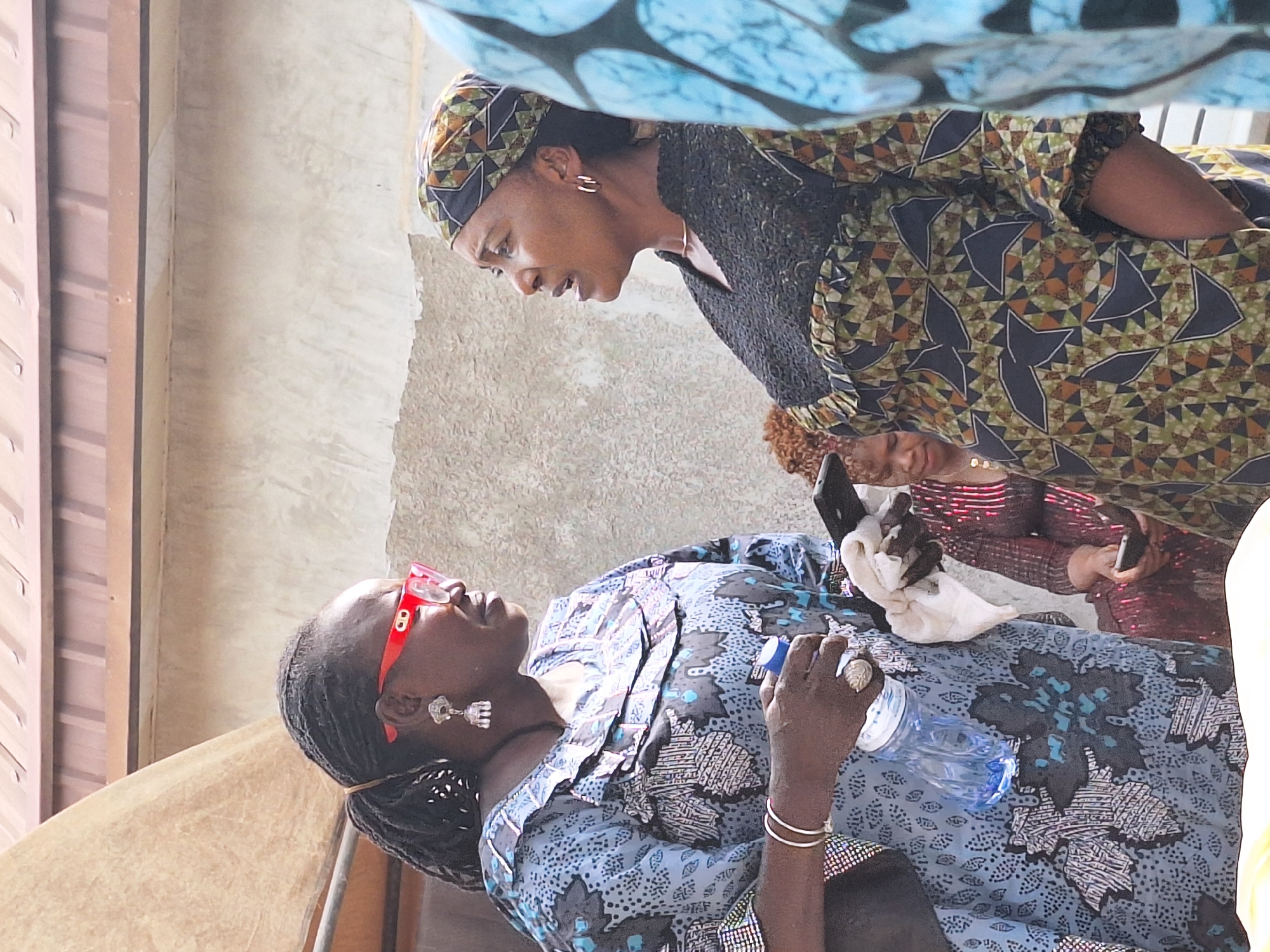 In Sabon Tasha, Nigeria, two women are interacting under a blue printed fabric. One woman stands wearing a headscarf and a patterned dress, looking down at the other woman who is lying down wearing red glasses and holding a water bottle. A third person is partially visible in the background, using a mobile phone. The scene includes a wall and what appears to be an outdoor or semi-outdoor setting.