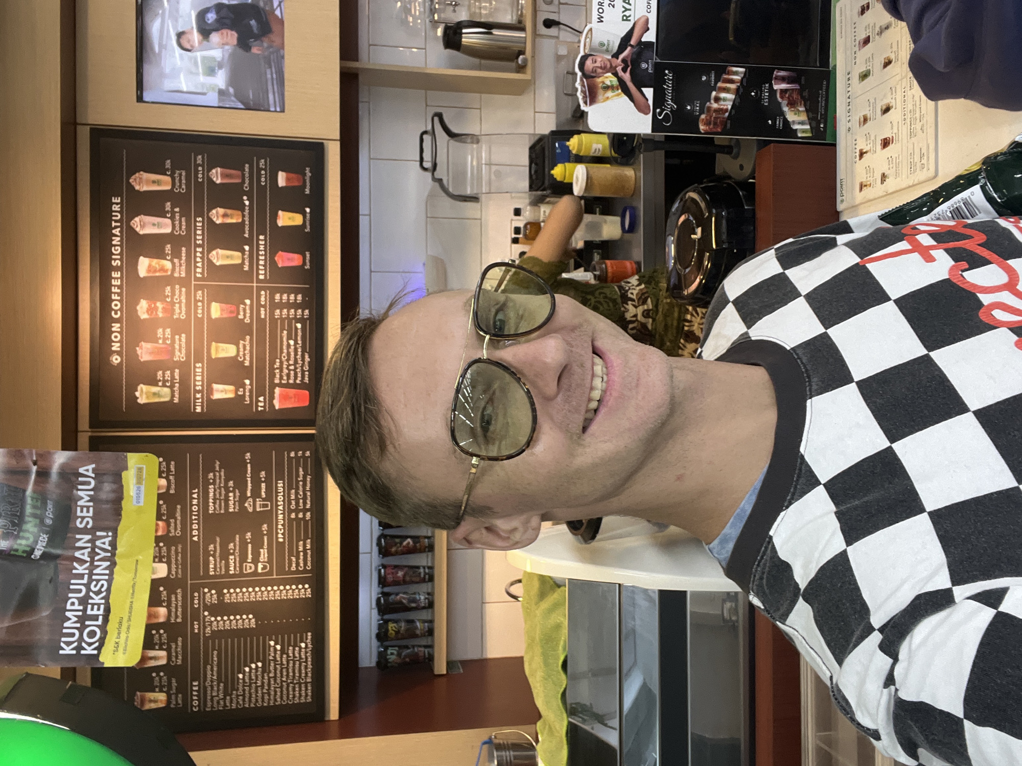 A man with short, light brown hair and a light complexion is taking a selfie in what appears to be a coffee shop. He is wearing a black and white checkered long-sleeved shirt and large, gold-rimmed sunglasses with yellow lenses. He is smiling. Behind him, there is a counter with various bottles and containers of syrups and sauces. To the right of the man, there is a menu board displaying coffee and non-coffee drink options, as well as a promotional poster featuring a man and the word "Signature." Further in the background, there are shelves with cups and a digital screen. The scene is indoors, with tiled walls and wooden accents. The establishment is likely a Point Coffee shop, given the branding visible on the menu and posters.