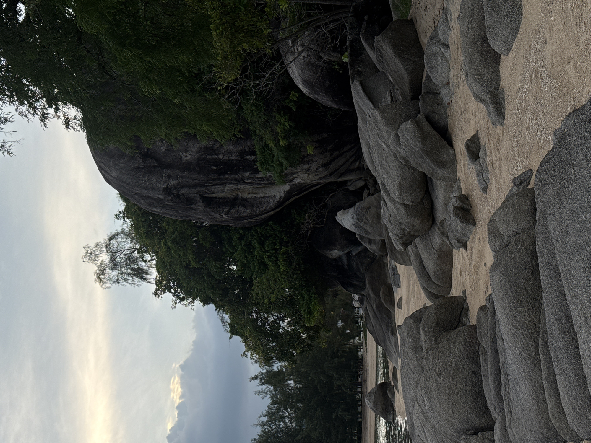 The image depicts a rocky beach scene, likely in Ban Bang Khao, Thailand, as suggested by the context. The foreground is dominated by numerous large, grey, rounded boulders scattered across a sandy beach. The sand appears to be a light beige color, with small white shell fragments visible in some areas. Beyond the boulders, the water's edge is visible, with gentle ripples suggesting calm water. In the mid-ground, a line of dark green trees forms a dense backdrop, with some taller, thinner trees interspersed further back. A massive, dark grey, irregularly shaped boulder is a prominent feature within the tree line, casting a shadow. The sky is overcast with clouds, with a faint glow of sunlight breaking through on the left side, indicating it might be early morning or late afternoon. There are no people or animals visible in the image. The overall atmosphere is serene and natural, emphasizing the geological formations and the coastal environment.
