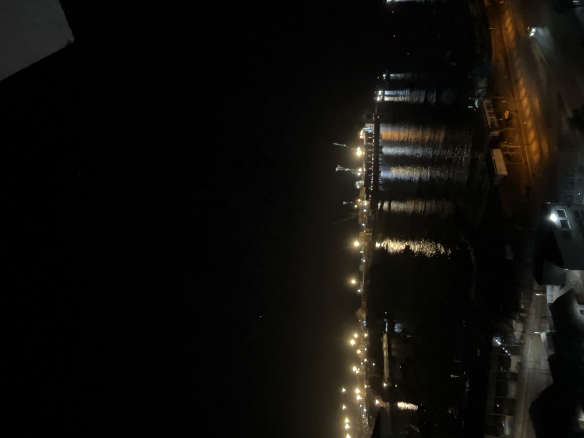 The image depicts a nighttime view of what appears to be a harbor or waterfront area. In the distance, lights indicate the presence of a structure, possibly a pier or an industrial complex, with reflections shimmering on the water's surface. A row of lights stretches along the coastline, marking a road or promenade. Closer to the viewer, buildings are faintly visible, suggesting an urban environment. The sky is dark, and the overall scene conveys a sense of tranquility and urban activity coexisting in the dark. The location is in the city of Unknown, Unknown.