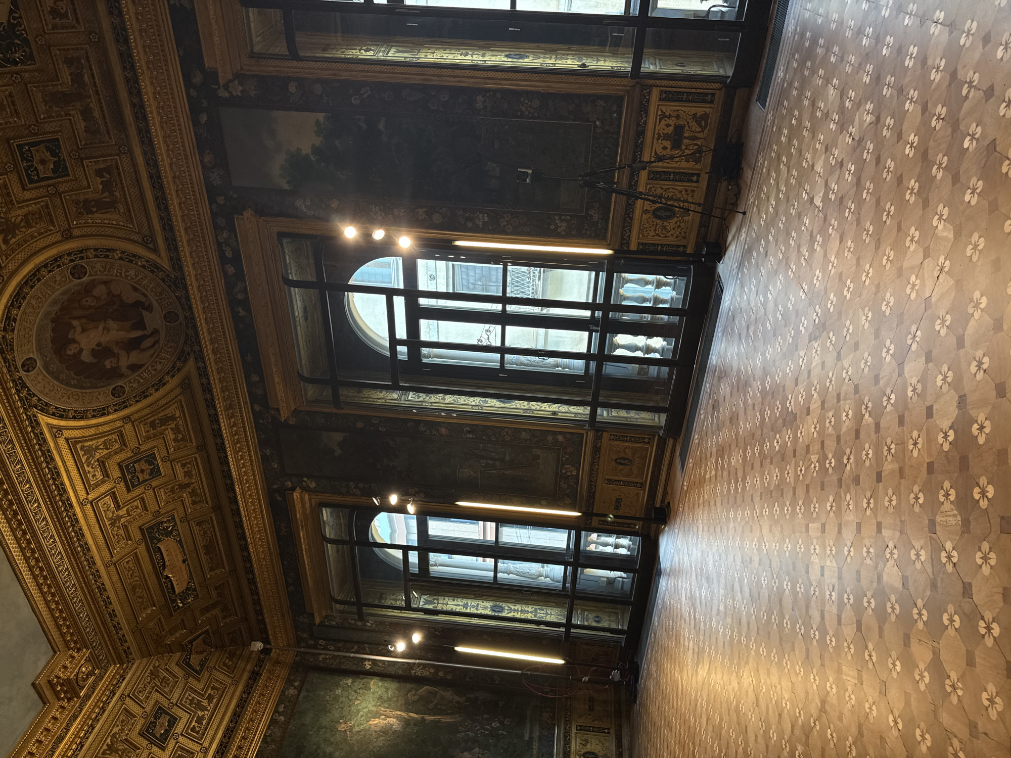 The image depicts the interior of a building in Milan, Italy, likely a historical or cultural site given the ornate architectural details.
The perspective is from below, looking upward. The ceiling is heavily decorated with gold trim, intricate carvings, and painted panels, including at least one with a circular medallion containing a painted scene with figures.
Two large windows with arched tops are visible. The windows have a black grid pattern and allow the viewer to see the building across the street. Between the windows are painted panels depicting landscapes. There is evidence of artificial lighting fixtures.
The floor is covered in tiles with a repeating floral pattern, giving the impression of an ornate mosaic.