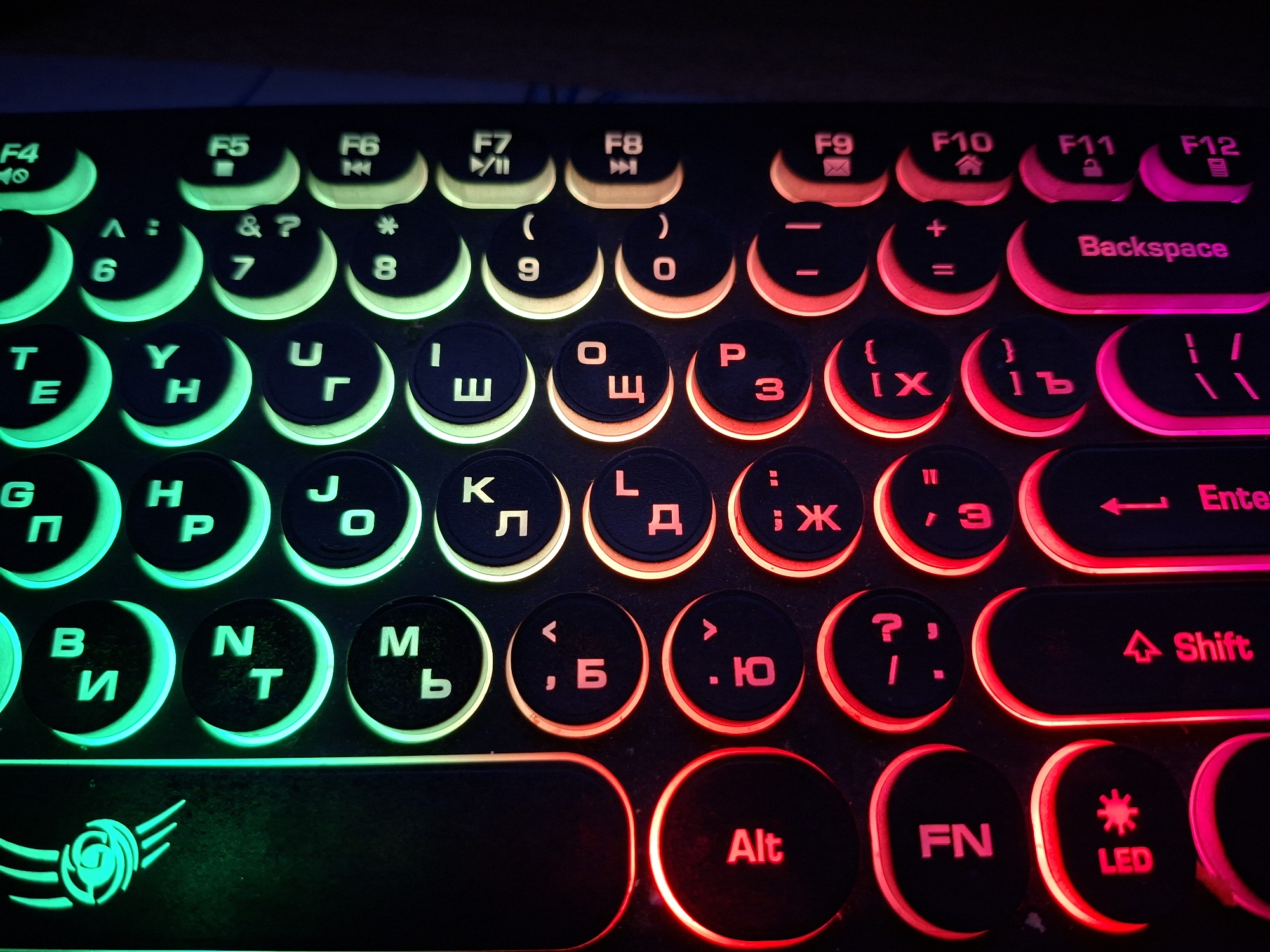 A close-up shot presents a gaming keyboard illuminated with vibrant LED backlighting. The lighting is multicolored, transitioning from green on the left to red and pink on the right, highlighting the keys' circular design and black bases. The keys are labeled with both English and Cyrillic characters, and function symbols, covering typical keyboard inputs, numbers, function keys (F1-F12), and special keys like 'Alt,' 'FN,' 'LED,' 'Shift,' 'Enter,' and 'Backspace.' The logo of the keyboard can be seen on the bottom left corner with a stylized design.