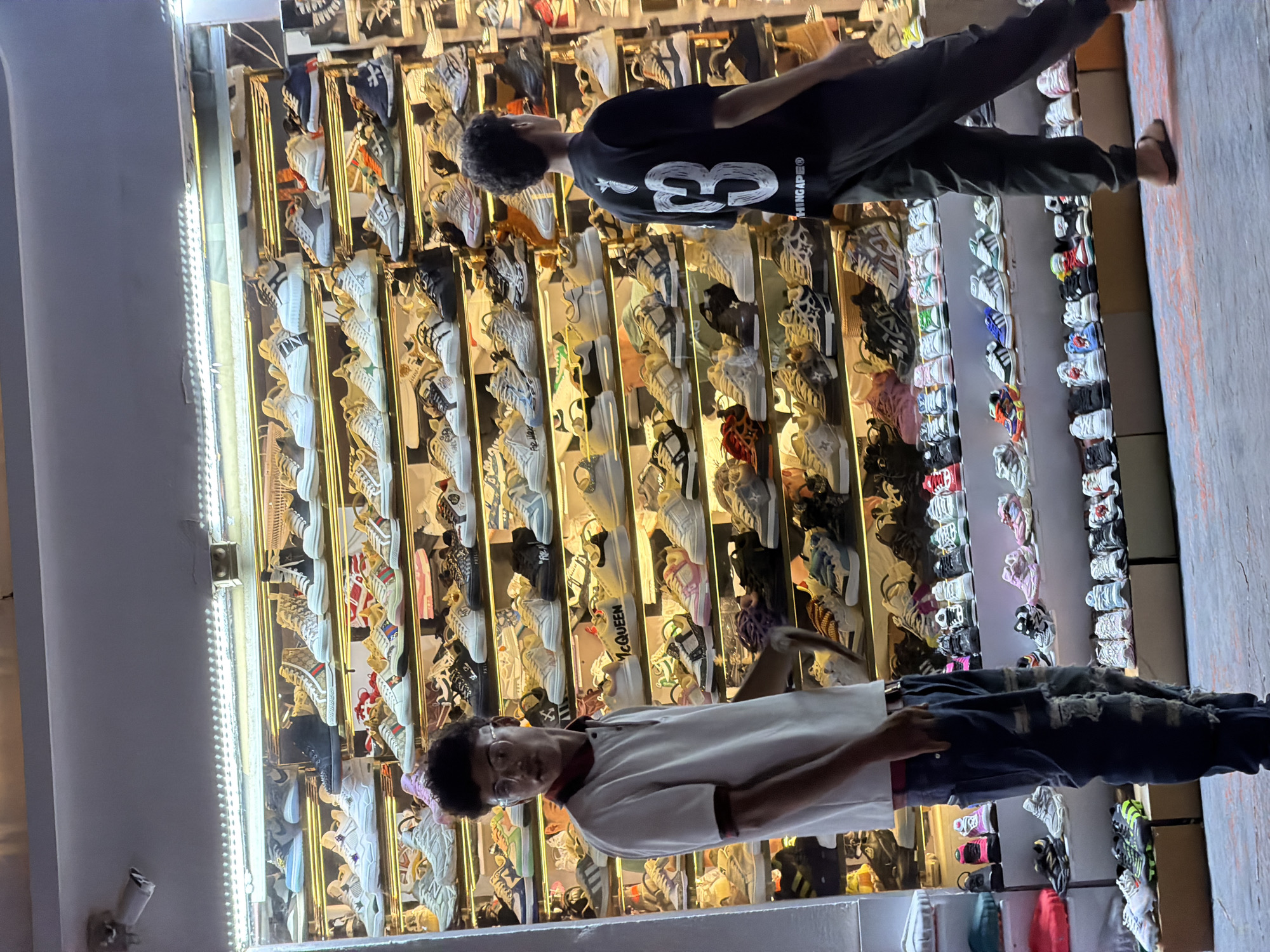 A dimly lit storefront displays a vast collection of athletic footwear on multiple shelves. The shoes, predominantly sneakers, are in a variety of colors and styles, suggesting a retail environment. Two young men are present. The one on the left, wearing a white polo shirt and distressed jeans, stands facing the camera, looking towards the footwear. The man on the right, in a black t-shirt and dark pants, is partially visible, facing away from the camera and towards the display. The lighting, primarily from fluorescent tubes above the shelves, creates bright highlights on the shoes and illuminates the scene. The background appears to be the interior of the shop, with more shelves and shoes receding into the darkness. The floor in front of the display is somewhat worn and shows faint colorful markings. The overall impression is a busy shoe store, likely in an urban area. The location is specified as Sharm El Sheikh, Egypt.