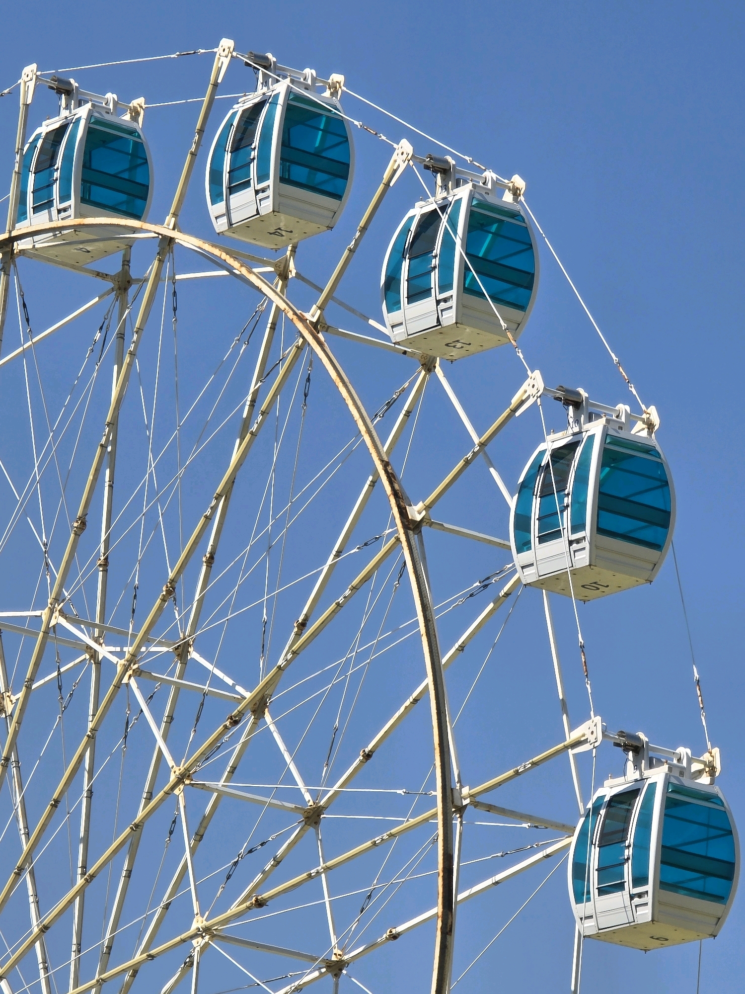 The image shows a Ferris wheel against a clear blue sky. Several gondolas, which are white with blue tinted windows, are visible attached to the wheel's structure. The Ferris wheel itself is made of a cream-colored metal framework with numerous supporting cables. The scene appears to be a sunny day, with no people or other discernible objects in the immediate vicinity. The location in the city of Polenitsa, Bulgaria, is not visually evident from the provided image.