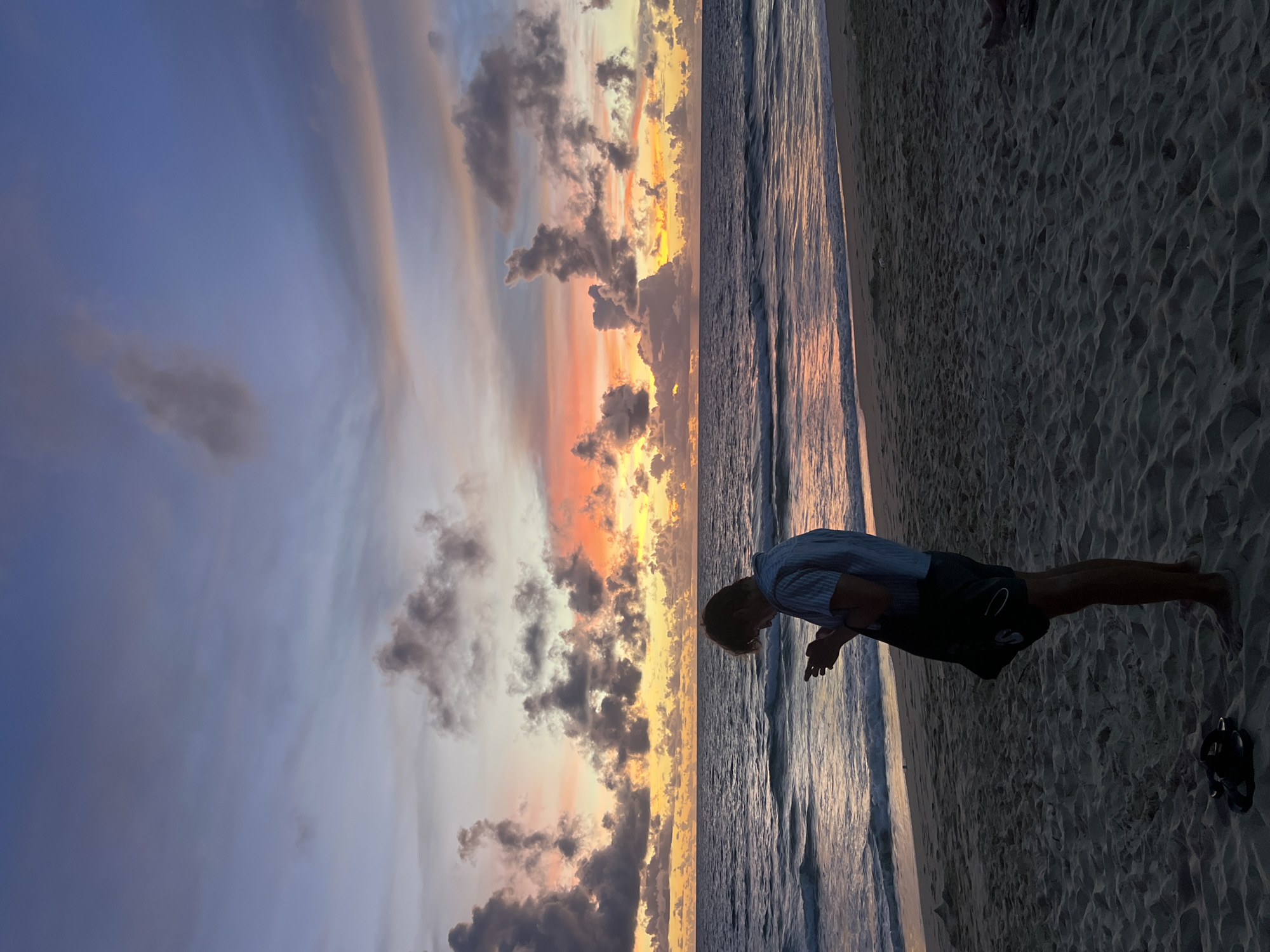 A person is standing on a sandy beach next to the ocean. The person is silhouetted against the sky and appears to be wearing a striped shirt and dark shorts. The person is bent over, with their hands clasped in front of them. The ocean is calm with small waves. The sky is filled with dramatic clouds in shades of orange, pink, and purple, indicating a sunset. The sand is light brown and textured, with footprints visible. The scene suggests a peaceful and serene moment at dusk. The location is Choeng Thale, Thailand.
