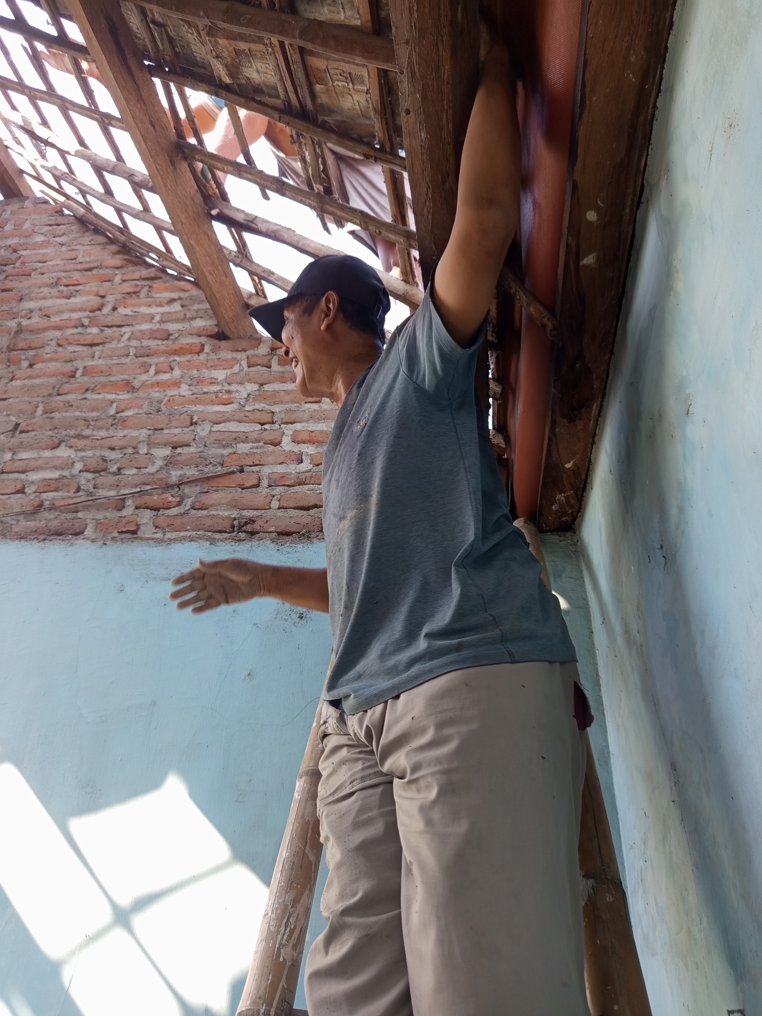 The image captures a man, appearing to be of middle age, standing on a bamboo ladder inside a building under renovation or construction, possibly located in Ngablak, Indonesia. The man is wearing a grey t-shirt, beige pants, and a dark baseball cap. He is facing to his left, with his mouth open as if speaking or shouting. His right arm is extended upwards, holding onto a wooden beam that is part of the roof structure. His left hand is outstretched in front of him, palm open, in a gesture. The background shows a partially constructed brick wall and a wooden roof frame with exposed beams and what appear to be roofing tiles. Light streams in from the damaged roof, casting shadows on a light blue wall. The scene suggests active construction or repair work is underway.
