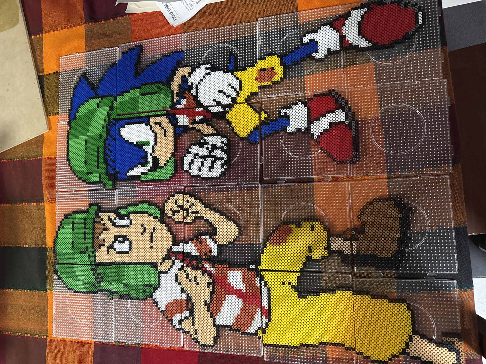 The image shows two pixel art figures constructed from Perler beads arranged on clear plastic pegboards. The top figure depicts Sonic the Hedgehog, recognizable by his blue quills, white gloves, yellow arms, and red and white shoes. The bottom figure appears to be King Hippo from the video game "Punch-Out!!" His figure is predominantly green, brown, white, red, and yellow. The figures are positioned on a tablecloth. Parts of a paper sheet with Spanish text are visible at the top left corner, as well as other objects, suggesting the presence of paperwork or documents. The context of the city of Puebla, Mexico, is not present in the visual information and would require external knowledge beyond what is provided.