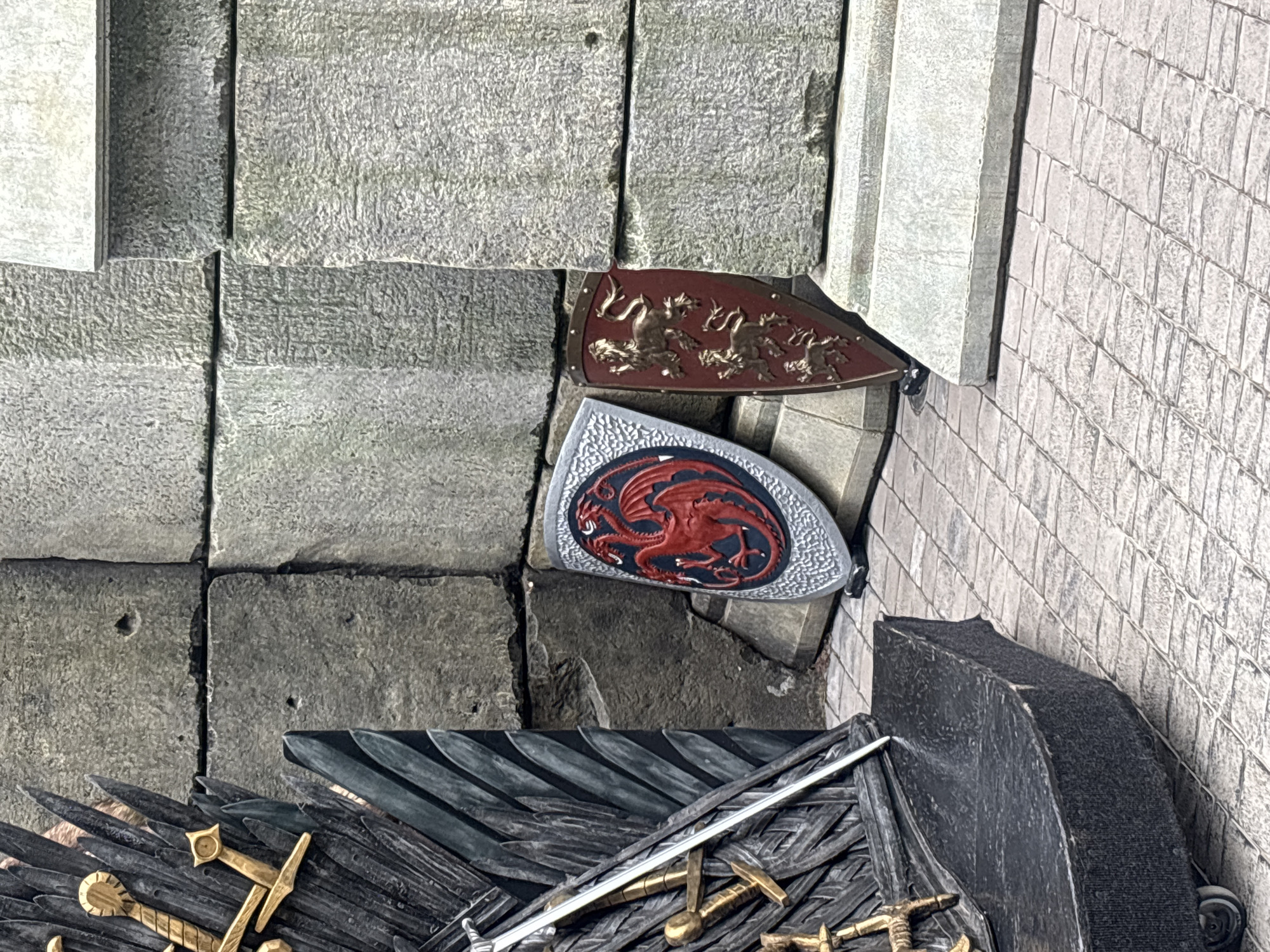 This is an outdoor, eye-level medium shot of two house sigils leaning against a large stone wall in Khryashchevka, Russia. The sigils are a shield with a red dragon on a black and silver background and a dark red shield with three golden lions. To the left is a portion of the Iron Throne, complete with swords and a sword resting on the throne. The ground is paved with square stones. The lighting is neutral.