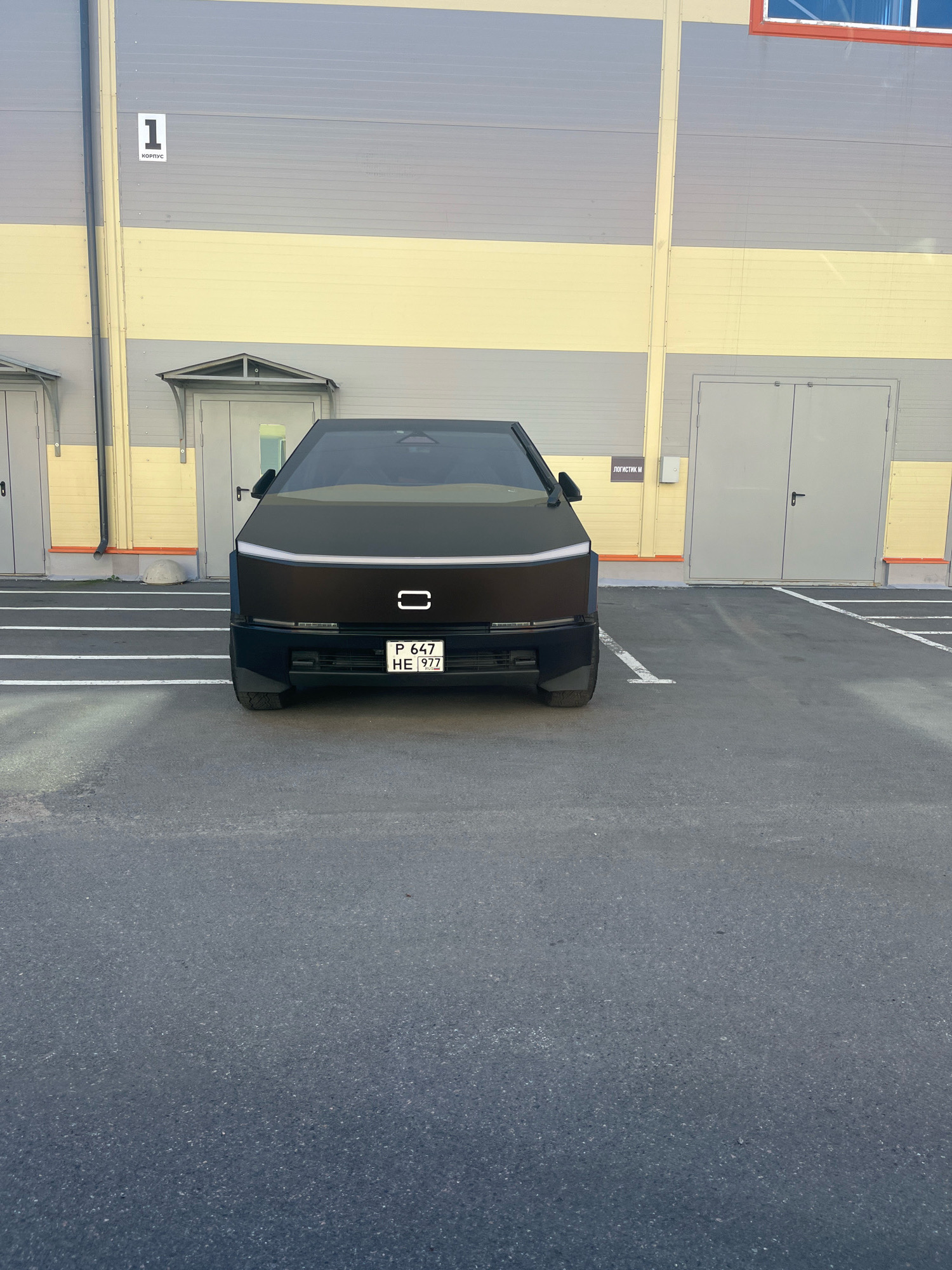A dark, angular vehicle, possibly a prototype electric truck, is parked in a paved lot outside a large industrial building. The building has horizontal bands of gray and yellow siding and several large, light gray doors. A sign on the building reads "1 корпус" (1 Building) in Russian. The vehicle has a distinctive illuminated front emblem and a license plate that reads "P 647 HE 977". White painted lines marking parking spaces are visible on the ground. The scene appears to be an industrial or logistics area in Saint Petersburg, Russia.