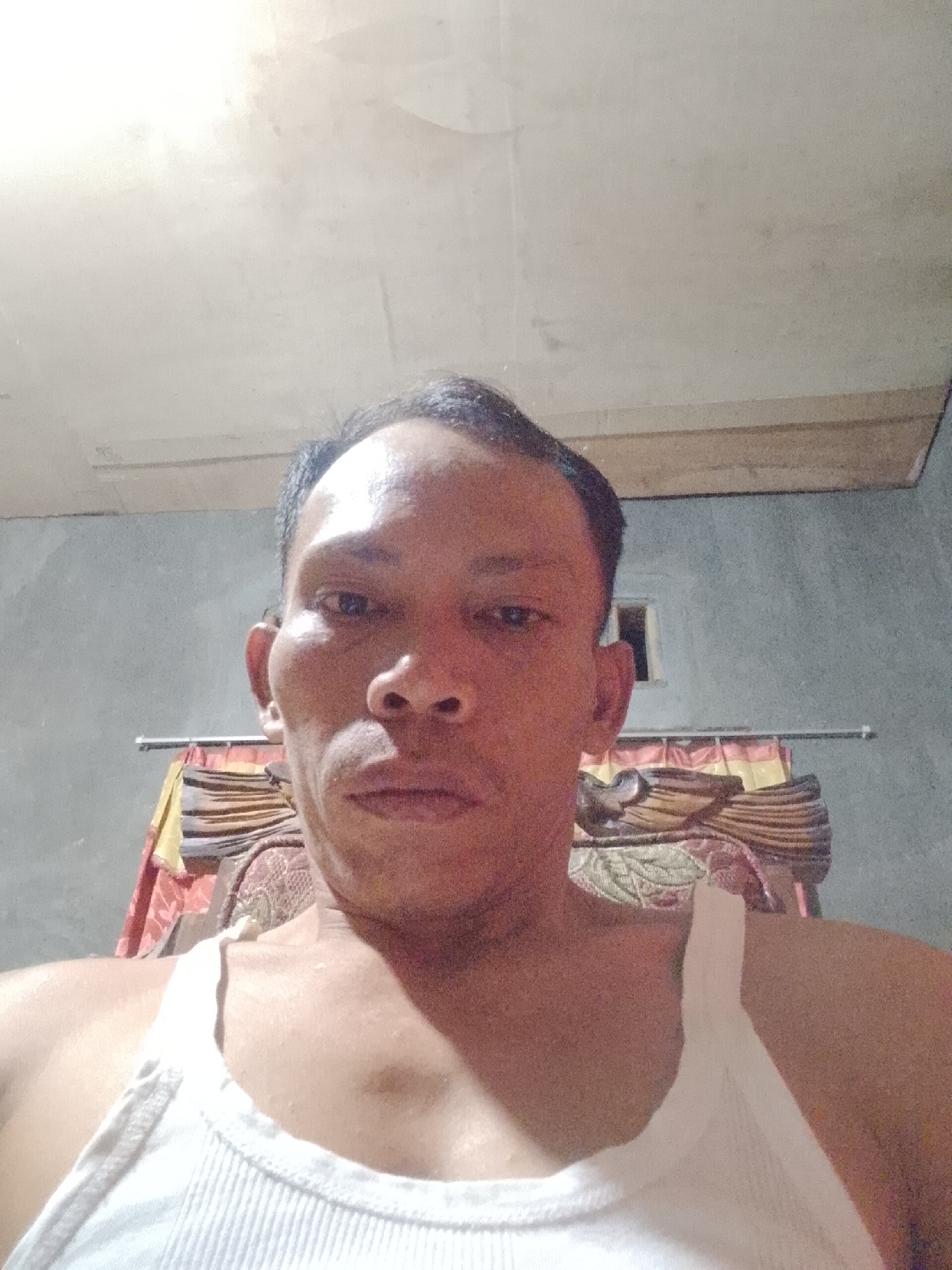 A man with dark hair and brown eyes, wearing a white tank top, looks directly at the camera. He is in a room with a gray concrete wall and a white ceiling. Behind him, a rack of clothing hangs on a horizontal bar. A small, dark-colored window is visible on the wall to his right. The image appears to be a selfie taken in Kali Cinta, Indonesia.