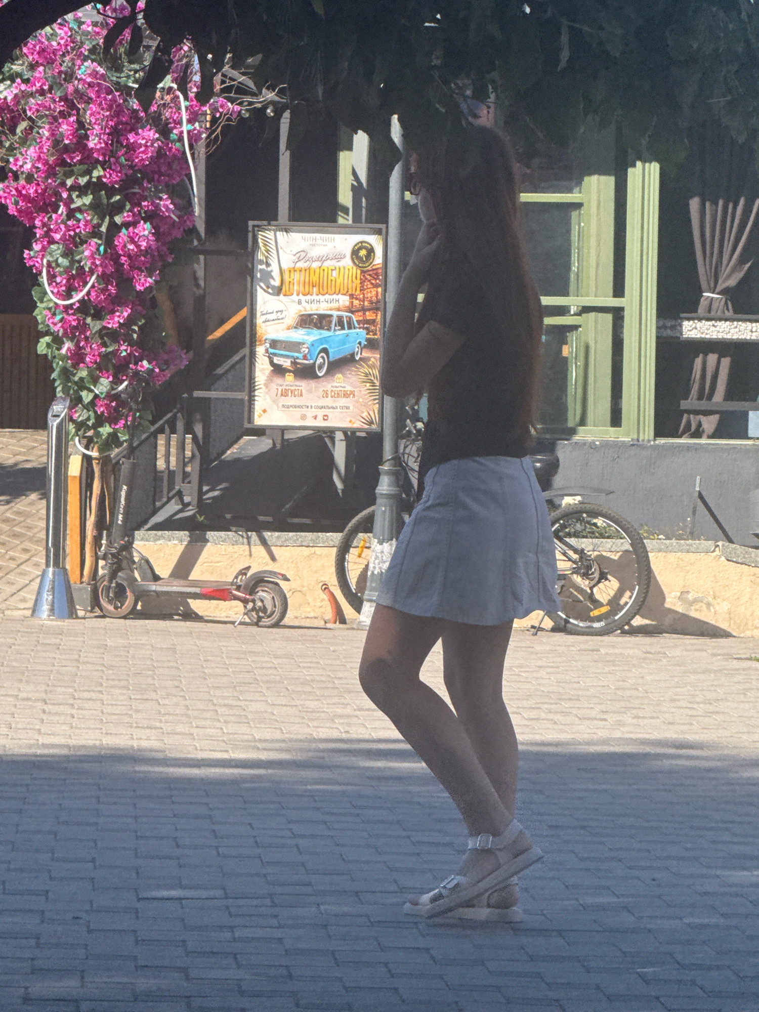 An Anapa, Russia scene depicts a young woman with long dark hair, wearing a dark t-shirt and a light blue skirt, walking on a paved sidewalk. She is wearing white sandals. To her left, there is a bright pink flowering bush. Behind her and to the left is a black electric scooter. Further in the background, a bicycle with a silver frame is leaning against a wall. A poster with an image of a blue vintage car and Russian text advertising "Avtomobiliya" and dates "7 Avgusta - 26 Sentyabrya" is displayed on a stand near the flowering bush. The environment appears to be outdoors, possibly a public area or street in Anapa, with sunlight casting shadows.