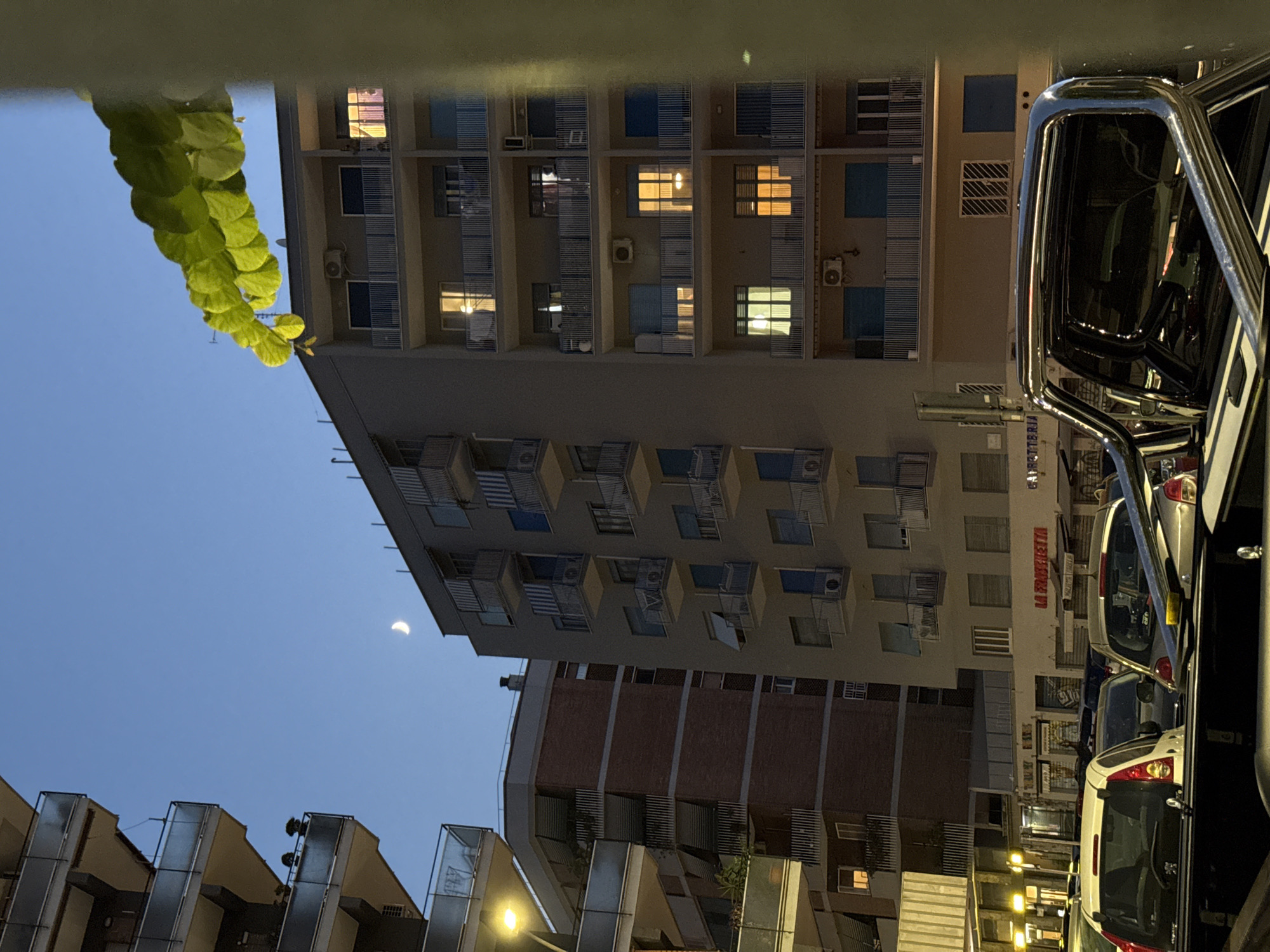 The image captures a twilight scene in an urban environment, likely Rome, Italy, with a clear sky and a visible crescent moon. In the foreground, a section of a modern pickup truck with a chrome roll bar is visible. Beyond the truck, multiple apartment buildings are depicted. The closest apartment building on the right is a multi-story structure with numerous balconies and windows, some of which are illuminated, indicating occupancy. To the left, another apartment building with a brick facade and balconies is shown. Further left, another residential building with balconies and a street lamp is visible. The lower part of the image shows parked cars and storefronts, including one with the signage "LA FRASCHETTERIA". The overall impression is of a residential street at dusk, with urban architecture and the natural element of the moon.