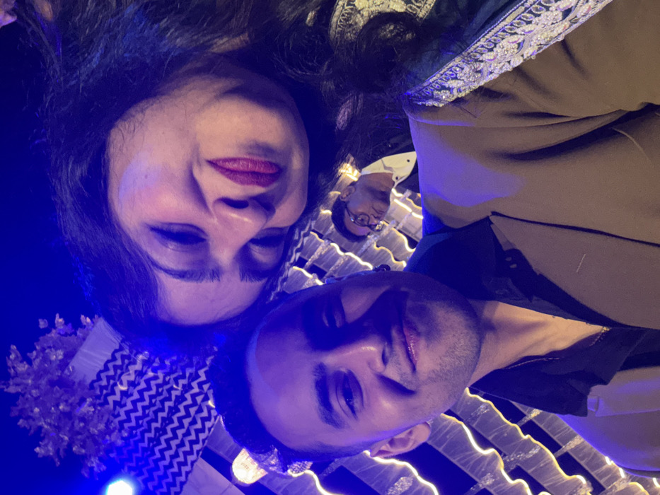 The image displays a close-up shot of several individuals illuminated by a striking blue light, giving the scene an atmospheric, cool tone. The image is oriented sideways, appearing rotated 90 degrees counter-clockwise from a typical upright perspective, suggesting it was taken from a low angle looking up at the subjects, or the camera was held at an unusual angle.
In the upper portion of the frame (left side in a typical upright view), a woman's face is prominently in focus. She has dark, wavy hair, defined eyebrows, and is wearing dark red lipstick, with a pleasant, slightly smiling expression. Her head is tilted slightly. Part of her shoulder and upper arm is visible, adorned with intricate white or silver embroidery on a dark fabric, suggesting a festive or formal garment.
Below and slightly to the right of the woman's face (center of the image in a typical upright view), a man's face is visible, also looking towards the camera. He has short dark hair and light stubble, and a faint, relaxed smile. He is wearing a dark top.
Further into the background, to the right and slightly below the first man (right side of the image in a typical upright view), another man's face is partially visible. He is wearing glasses and a white collared shirt with a dark outer garment, possibly a suit jacket. This individual is less in focus compared to the two in the foreground.
The entire scene is bathed in an intense blue light, which also casts a purplish hue on parts of the individuals' faces and clothing, creating a distinct mood, possibly for an evening event or a venue with specialized lighting. In the background and around the subjects, luminous patterns are visible, likely from decorative lighting installations such as string lights or LED strips, forming wavy or zigzag lines. To the bottom left of the original orientation, there's a hint of a lighter, possibly floral or textured fabric decoration.
Overall, the image captures a social moment, likely a group selfie or photo, at an event like a party, club, or themed gathering, characterized by its unique blue lighting and the relaxed, pleasant expressions of the subjects.