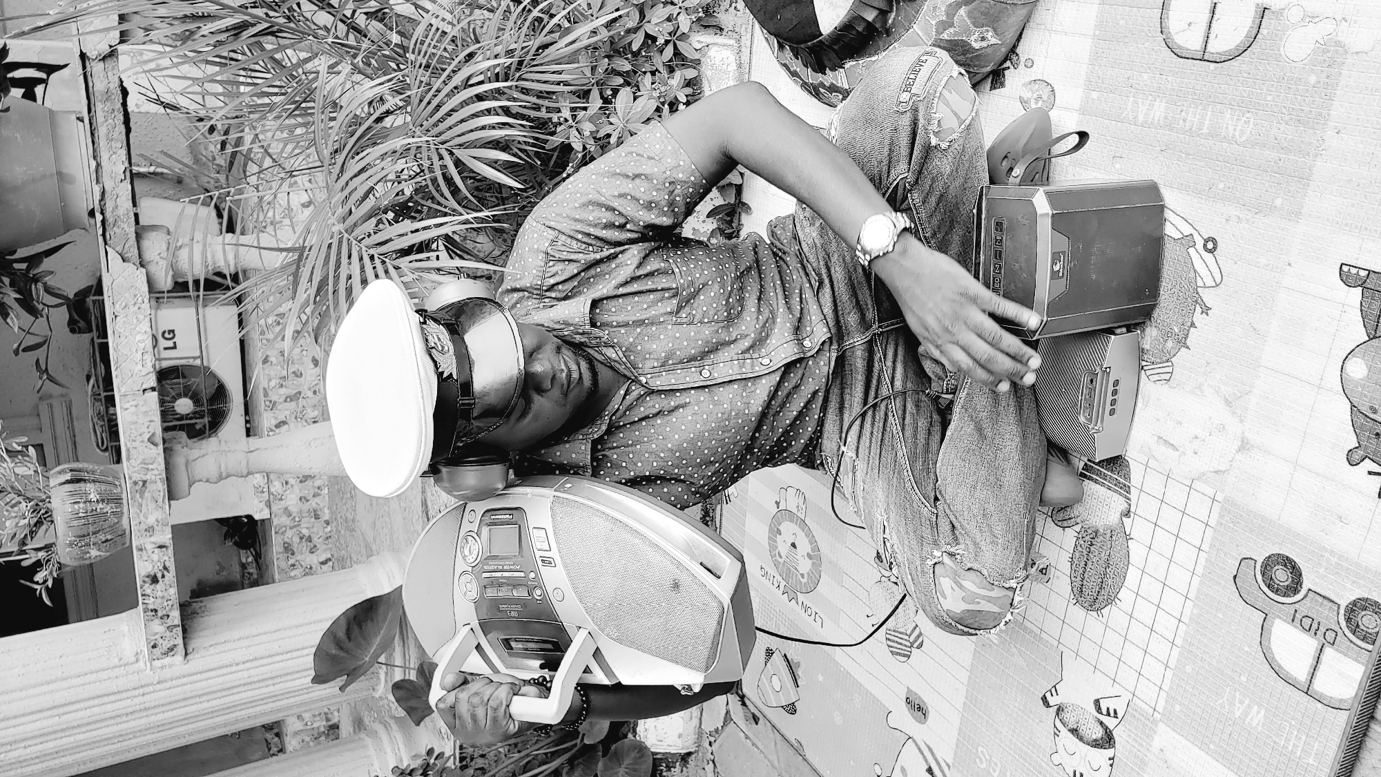 A black and white, medium shot shows a man sitting cross-legged on a patterned mat outdoors. He is wearing a patterned short-sleeved shirt, ripped denim jeans, a white cap, and headphones. He holds a boombox in his left hand and rests his right hand on a smaller speaker. The man is looking down at the speaker. Behind him, a palm plant and a large building with columns are visible. An air conditioner with the LG logo is mounted on the building wall. The scene appears to be in Narayi, Nigeria.