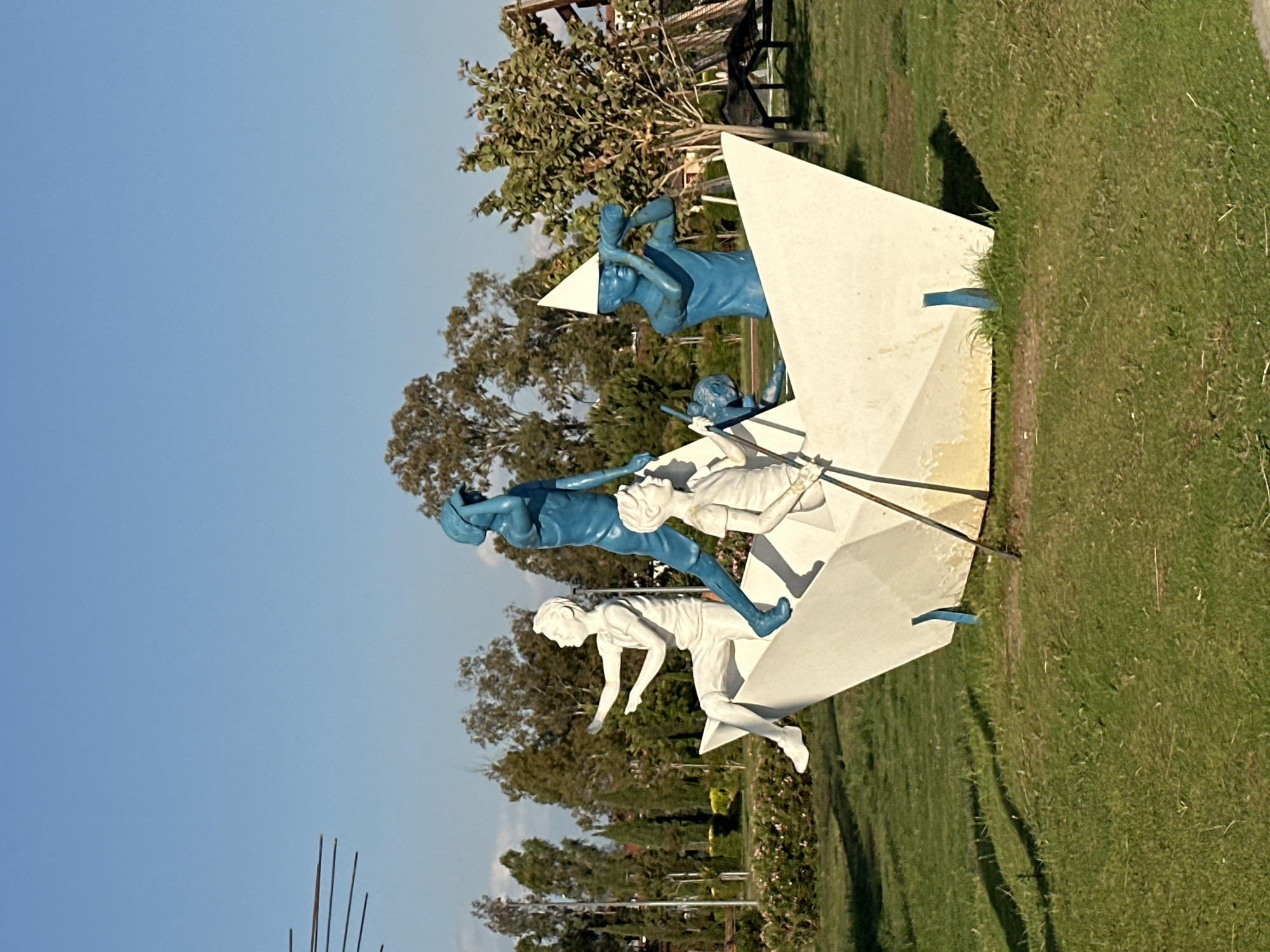 The image depicts a modern art sculpture situated in a green, grassy area, presumably a park or open space. The sky above is a clear, light blue. The sculpture consists of abstract white geometric shapes supporting figures in motion, some painted white and others painted a distinct blue.
Located in Fethiye, Turkey, the specific location is identified as the "Sehit Fethi Bey Parki" or "Fethiye Martyrs Memorial Park". The park features the 'Sporting Activities of Fethiye' sculpture, which is depicted in the image. The sculpture appears to depict individuals participating in various sporting activities.