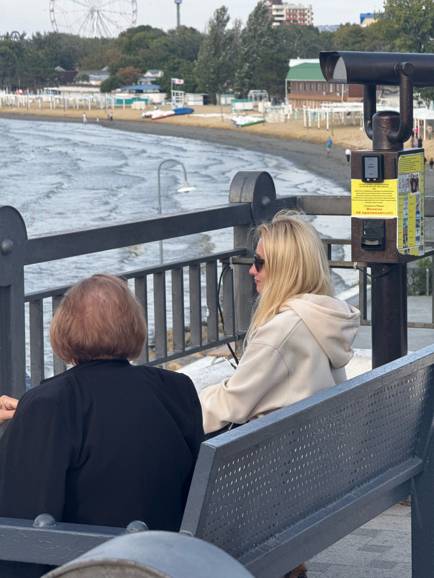 In Anapa, Russia, two individuals are seated on a bench facing a body of water. On the left, a person with short, reddish-brown hair is wearing a dark jacket. To their right, a blonde woman with sunglasses is dressed in a light-colored hooded sweatshirt. Behind them, a large viewing scope is mounted on a pole. The scope has a sign with Russian text and what appears to be a coin slot. The background shows a beach with umbrellas, buildings, and a Ferris wheel. The water in front of the beach is choppy.