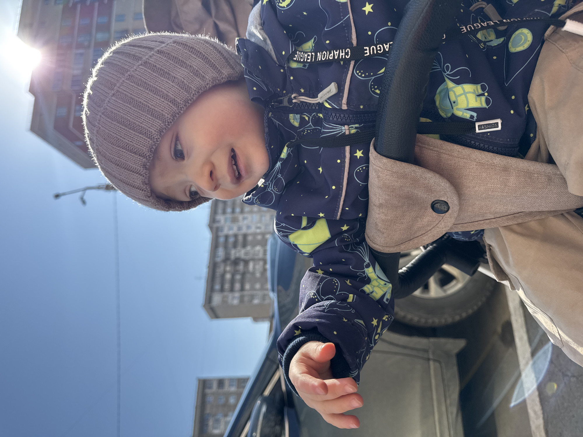 A toddler wearing a brown knit hat and a blue patterned jacket is seated in a stroller. The jacket has a zipper and the words "CHAMPION LEAGUE" printed on it. The stroller has a brown fabric attachment and a black handle. The child is holding their hand out. In the background are buildings, a bright sun, and a blue sky. The photo appears to be taken at an angle. Based on the architectural style and urban layout, the location is likely Izhevsk, Russia.