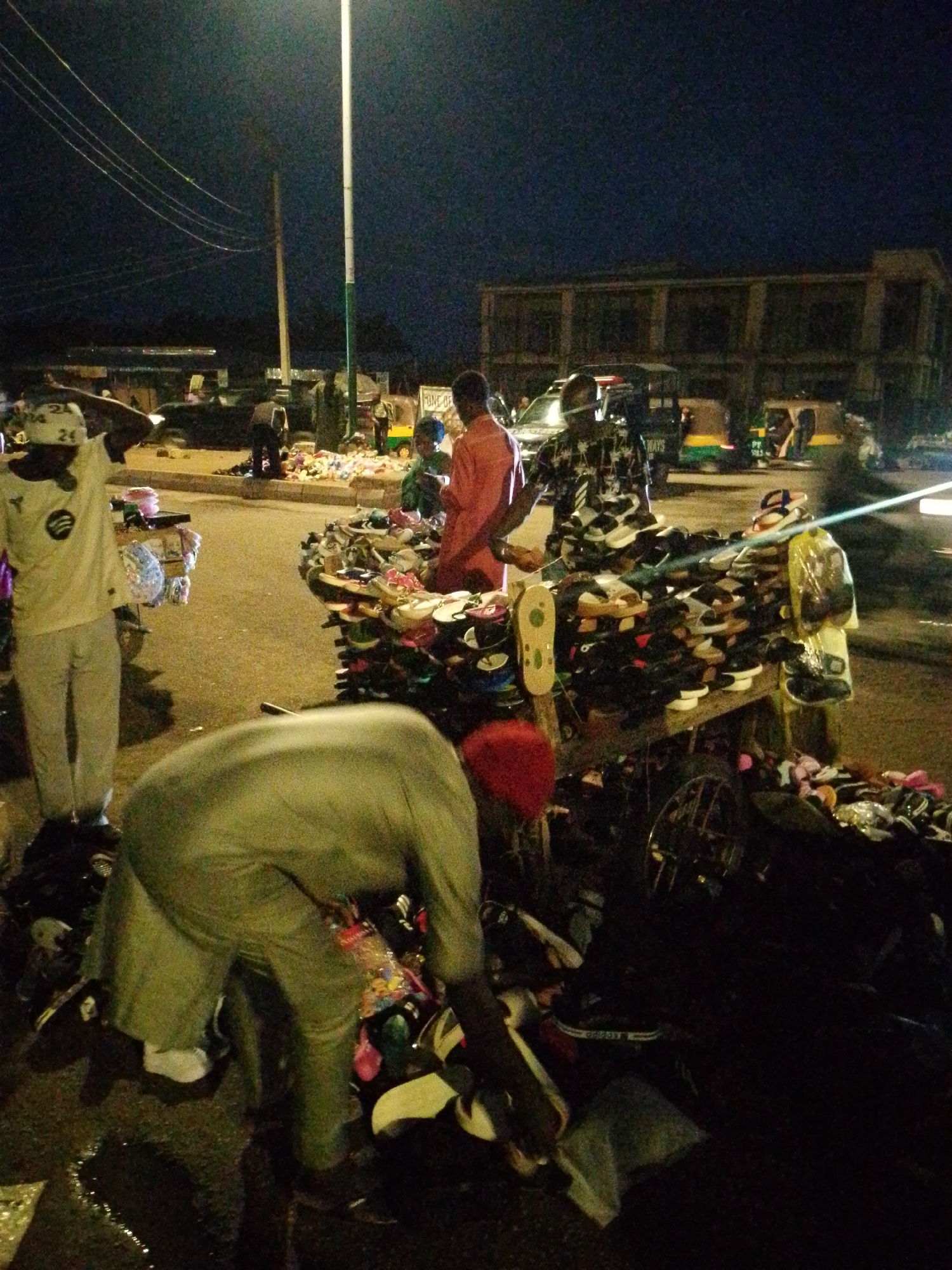 This is a nighttime image of an outdoor market, likely in a city. The scene is dimly lit, with artificial lighting from streetlights and possibly nearby buildings. In the foreground, a person wearing a light-colored shirt and pants is bent over, appearing to sort through items on the ground. To the left, another person in a white shirt with a logo and light-colored pants is standing, with one hand on their head.
Dominating the mid-ground is a display of shoes, predominantly sandals and flip-flops, arranged on a cart and on the ground. Several people are visible behind the shoe display, including one person in an orange garment and another in a patterned shirt. In the background, there are vehicles, including what appear to be auto-rickshaws and possibly a car. A multi-story building with a colonnaded facade is visible in the distance, and utility wires are strung across the dark sky. The ground appears to be paved asphalt, and there are scattered items and reflections on the wet surface in the lower left.