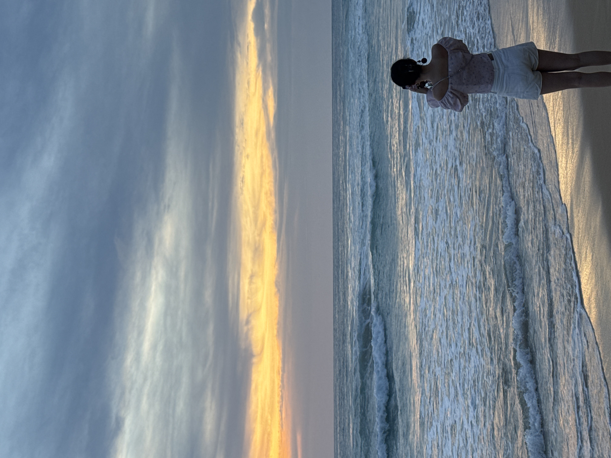 The image shows a person standing on a beach in Karon, Thailand, looking out at the ocean. The person is wearing a pink off-the-shoulder top and white shorts. They are holding a phone up to take a picture. The sky is cloudy with hints of orange and yellow from a sunset. The ocean has waves rolling in. The sand is wet where the waves have receded.