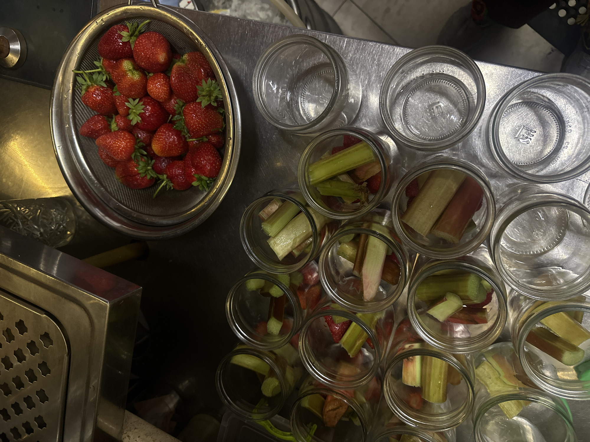 A top-down, slightly angled medium shot captures a kitchen preparation scene with strawberries and rhubarb in glass jars. A stainless steel colander overflowing with fresh strawberries sits in the upper right. Below, numerous clear glass jars are arranged, many filled with cut pieces of rhubarb and some with diced strawberries. Some jars are empty, revealing their textured bases. The workspace appears to be a metal countertop. The background shows a stainless steel preparation surface with a perforated pattern. The overall lighting is dim and appears to be indoor artificial light. There are no people visible in the image. There is no information to suggest this image is located in Ternopil, Ukraine.