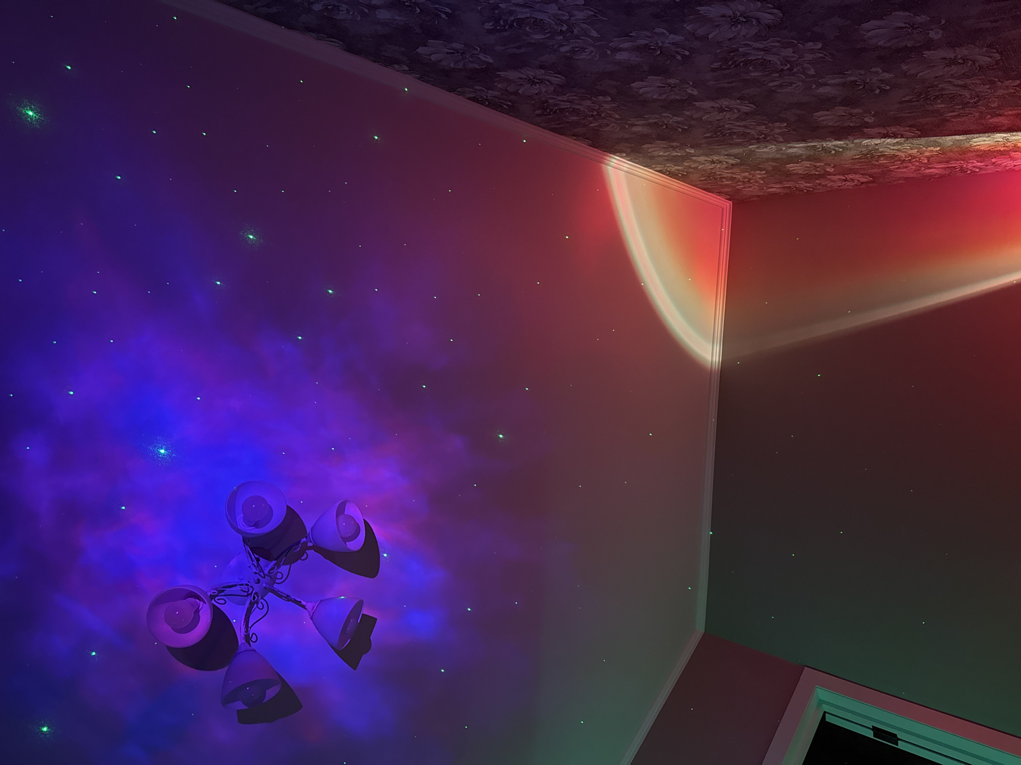 The image shows the interior of a room, likely a bedroom, with a ceiling fan and a patterned wallpapered wall. The ceiling is illuminated with a nebula-like projection in shades of purple and blue, with numerous small green dots resembling stars scattered across it. A segment of a projected rainbow or sunset light arc is visible on the ceiling and wall in the lower right portion of the image. The room appears to be unoccupied.