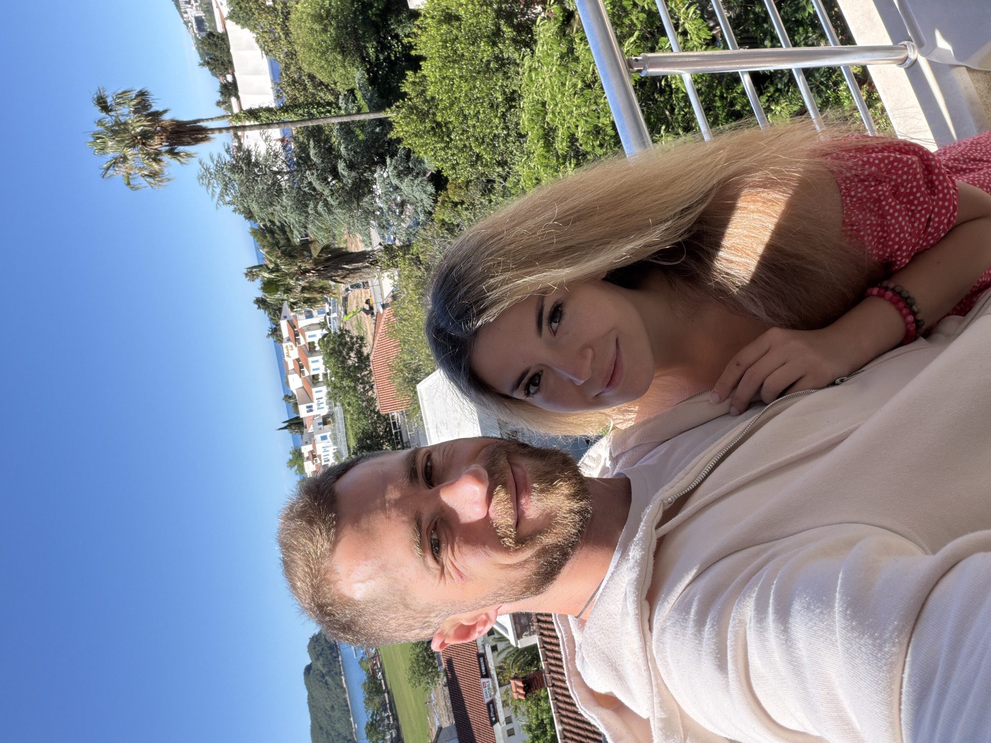 A photo shows a man and a woman, presumably in the city of Budva, Montenegro. The man, closer to the camera, has a beard and is wearing a light-colored hooded sweatshirt. The woman has long blond hair and is wearing a red polka-dot dress. They are likely on a balcony overlooking a landscape with buildings, trees, and a view of the sea. A palm tree is visible in the background. The sky is clear and blue, indicating a sunny day. The scene is a casual, possibly spontaneous, photograph taken by one of the subjects.