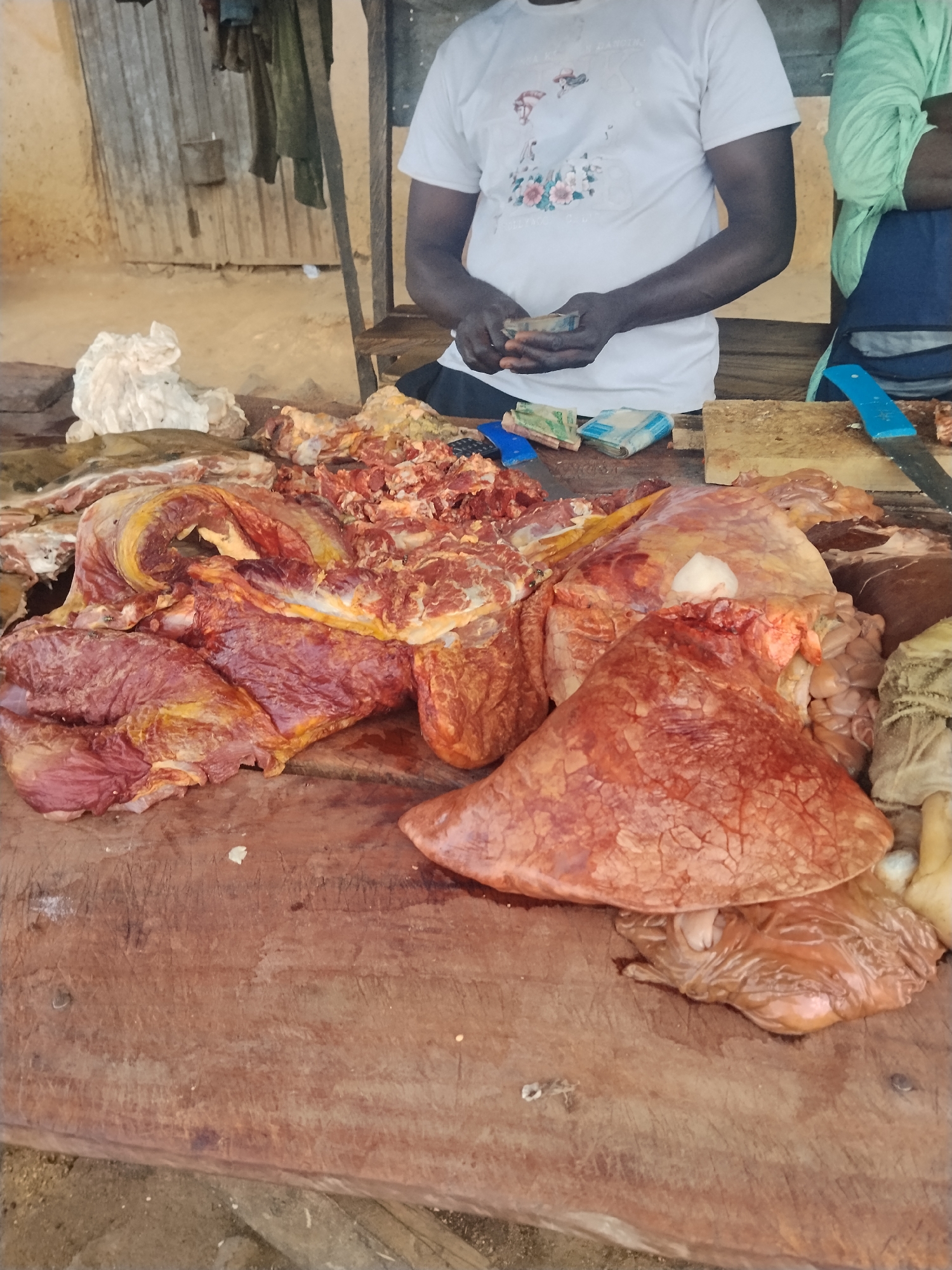 A person wearing a white t-shirt and black shorts is standing in a market stall in Mangu Town, Nigeria, holding a bundle of Nigerian currency. The stall is filled with raw meat, including large cuts of red meat and what appears to be internal organs. A person in a light green shirt and dark blue apron stands to the right of the frame. A wooden cutting board with knives and more raw meat is in the foreground. The background consists of corrugated metal sheeting and wooden planks, indicative of a rustic market environment.