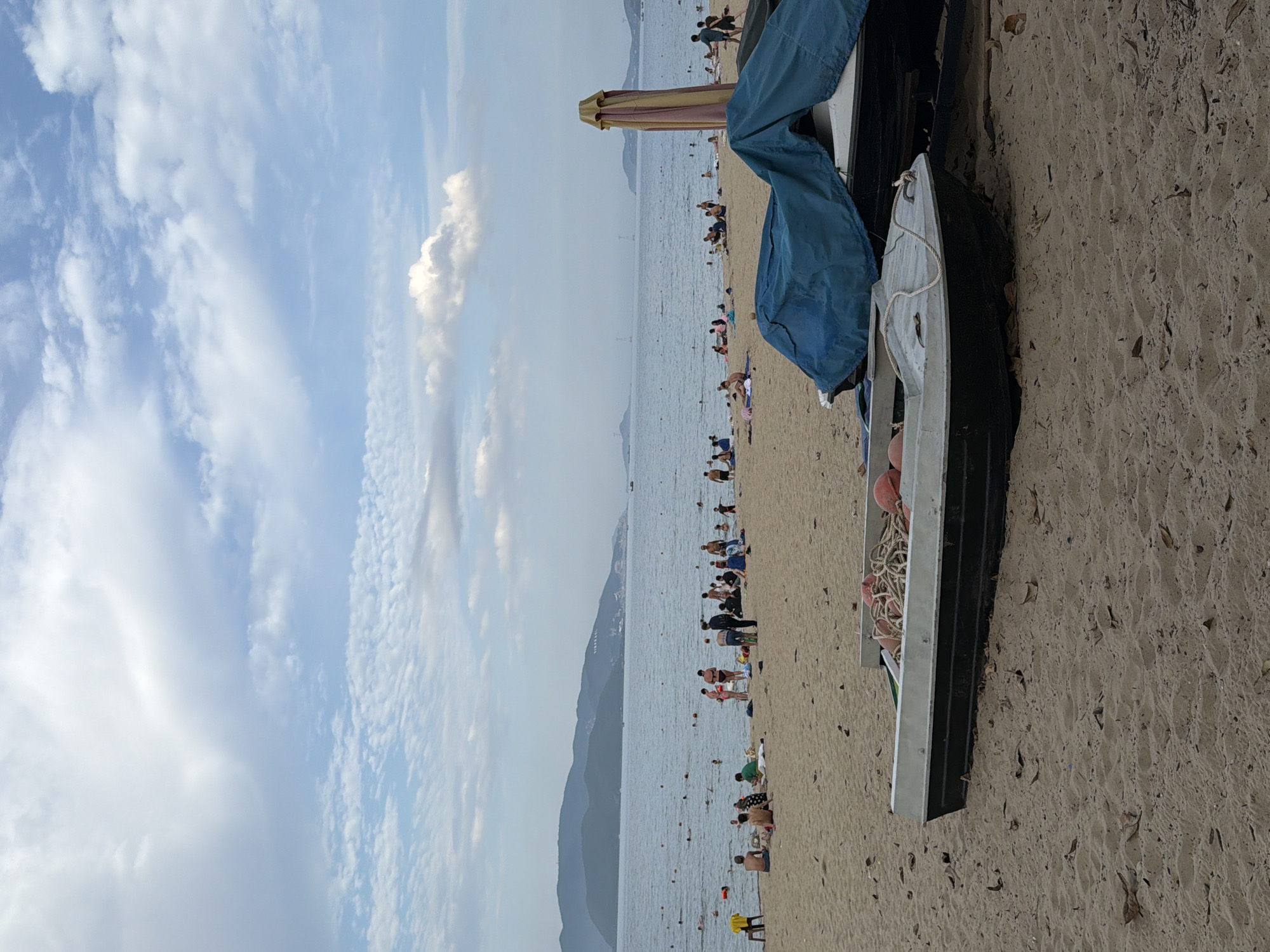 The image depicts a wide, sandy beach in Nha Trang, Vietnam, under a partly cloudy sky. The sea is calm and stretches towards a hazy coastline with distant mountains. The beach is populated by numerous individuals engaged in various activities. Some are swimming or wading in the shallow water, while others are sunbathing or sitting on towels and blankets on the sand. There are several boats pulled up onto the beach, some covered with blue tarps, and others with ropes and buoys visible. A large, closed umbrella with a striped canopy stands on the sand. The overall atmosphere appears relaxed and recreational, typical of a popular beach destination.