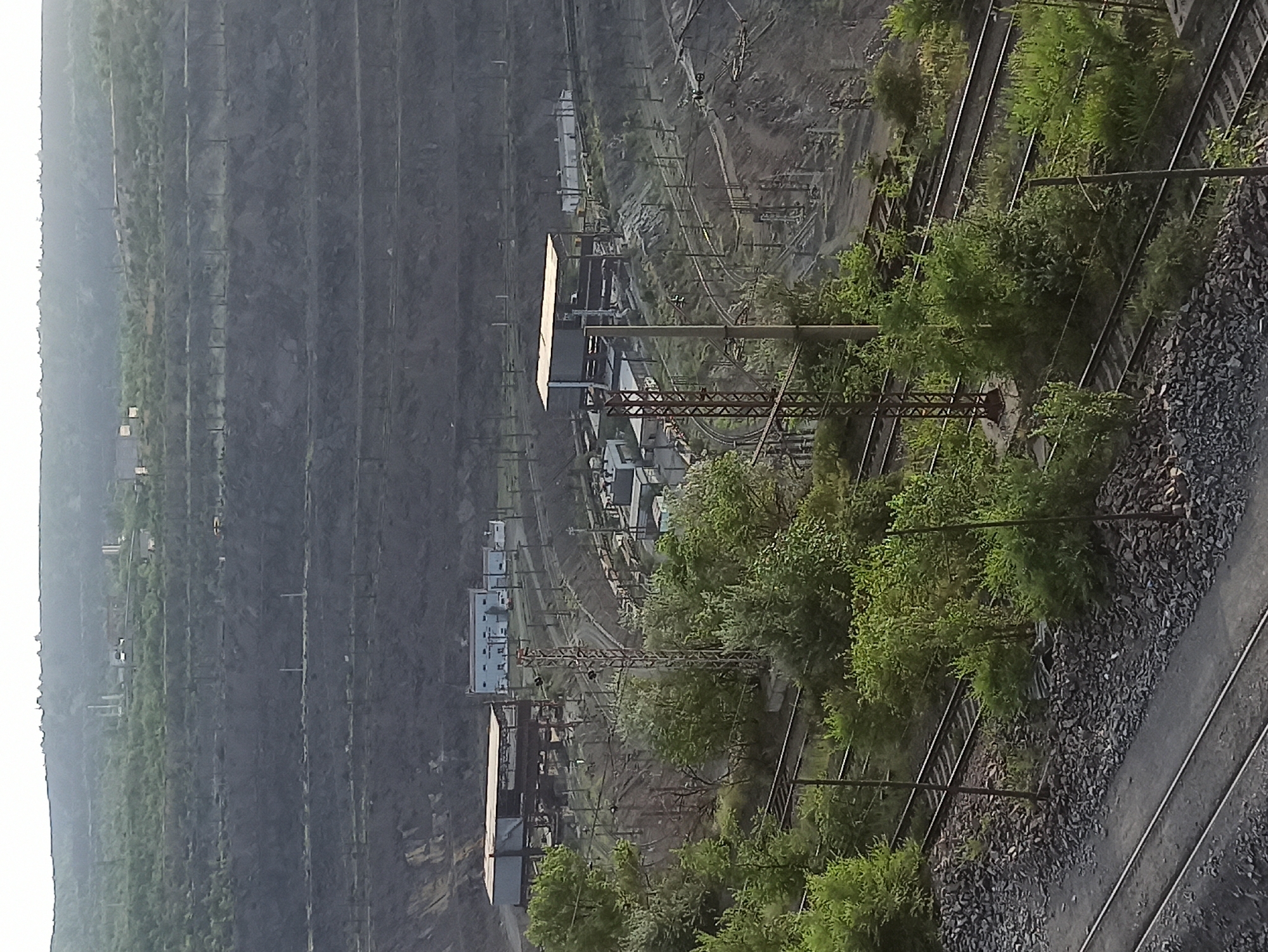 The image shows a complex industrial or transportation hub set against a backdrop of steep, terraced hillsides. The primary focus is on a network of train tracks, with multiple parallel lines weaving through the landscape. Along the tracks, there are various structures, including what appear to be warehouses or industrial buildings, and what look like electrical substations or railway signaling equipment. Overhead power lines and support structures are prominent, suggesting an electrified railway system.
Greenery, in the form of trees and bushes, is scattered throughout the scene, with some areas showing dense foliage crowding the tracks. The hillsides are largely barren and rocky, with defined terracing, possibly from mining or construction. The lighting suggests it is daytime, possibly late afternoon or early morning, as the light appears somewhat soft and diffuse. There are no people visible in the image, and the overall impression is one of organized, large-scale infrastructure. The scene is static, depicting a functional, operational area. There is no visible text in the image.