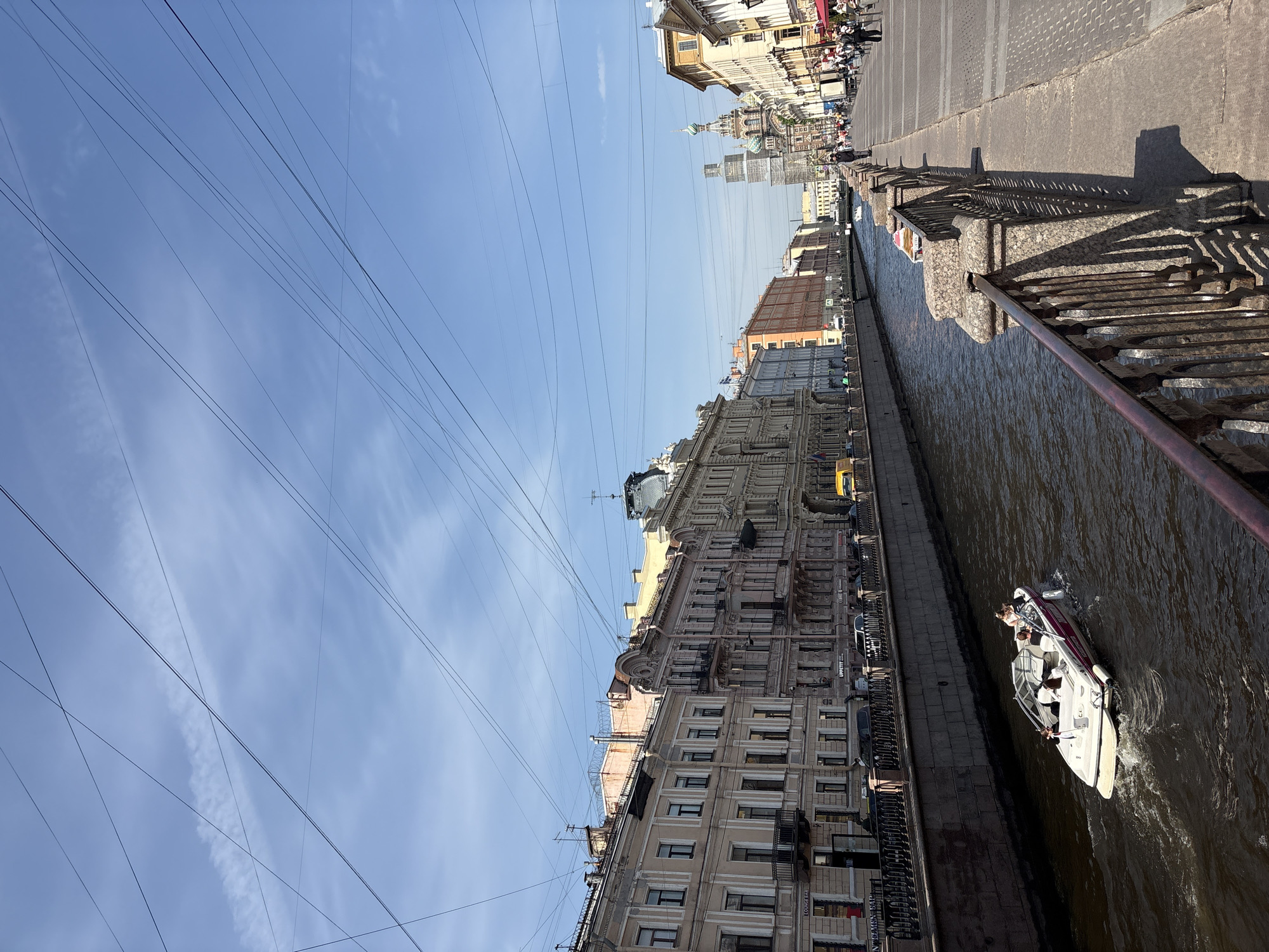 The image captures a view of a canal in Saint Petersburg, Russia. A motorboat with several people on board travels through the water. The canal is lined with buildings featuring classical architecture. Above, numerous power lines crisscross the sky. A sidewalk with a metal railing borders the canal. Pedestrians are visible on the sidewalk in the distance.