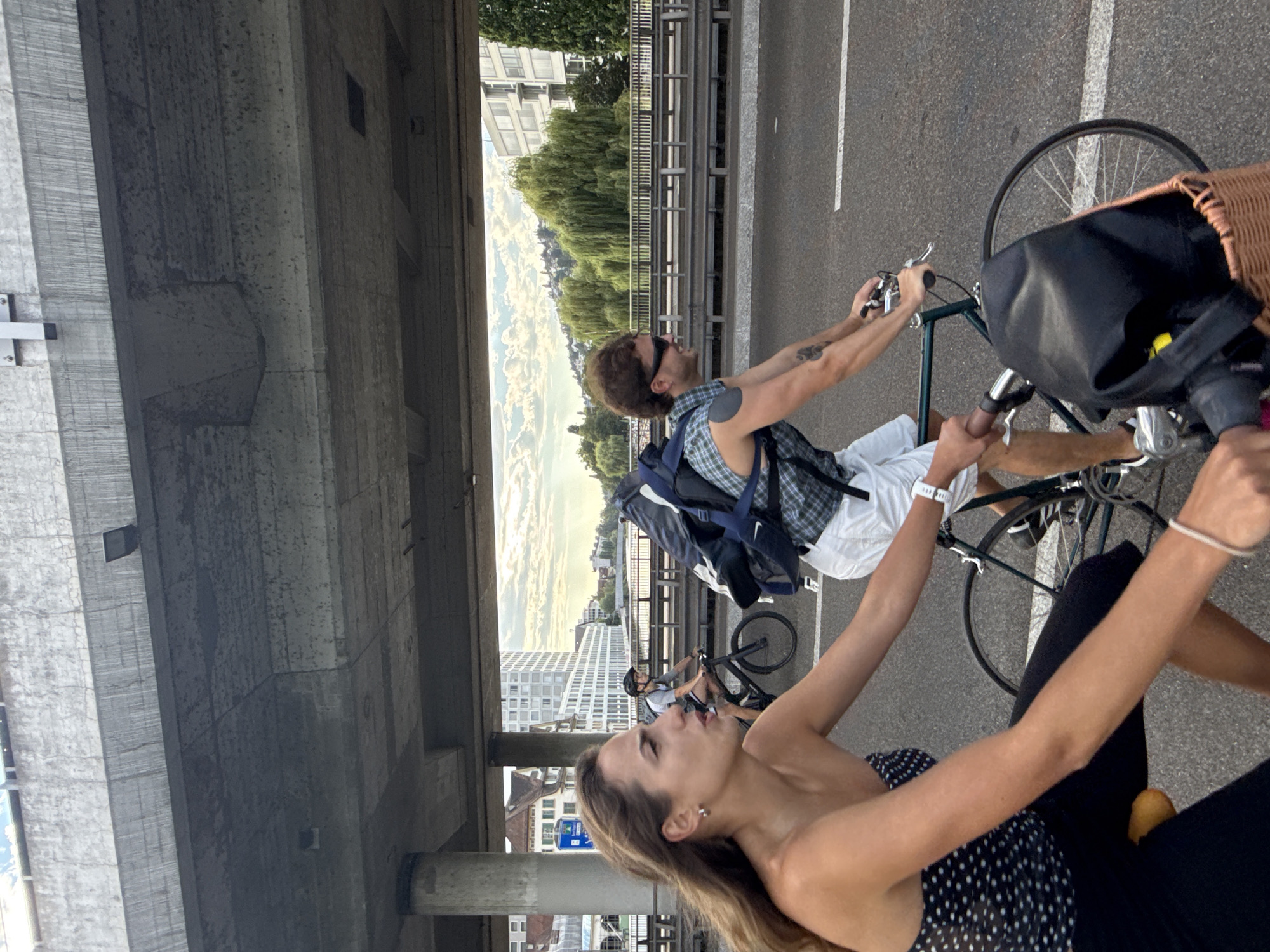 The image captures a street scene in Zurich, Switzerland, under a concrete overpass. In the foreground, a woman with long brown hair rides a bicycle, wearing a black and white polka dot top and black pants. She is looking to her left, with a focused expression. Behind her, a man with brown hair, sunglasses, and a plaid shirt with white shorts also rides a bicycle. He has a large backpack on and is looking to his right, smiling. Another person on a bicycle with a helmet is visible further in the background. The background reveals a cityscape with buildings, trees, and a river, suggesting a bridge or elevated roadway. The lighting indicates it is daytime, possibly late afternoon. The overall atmosphere is one of everyday urban life and activity.