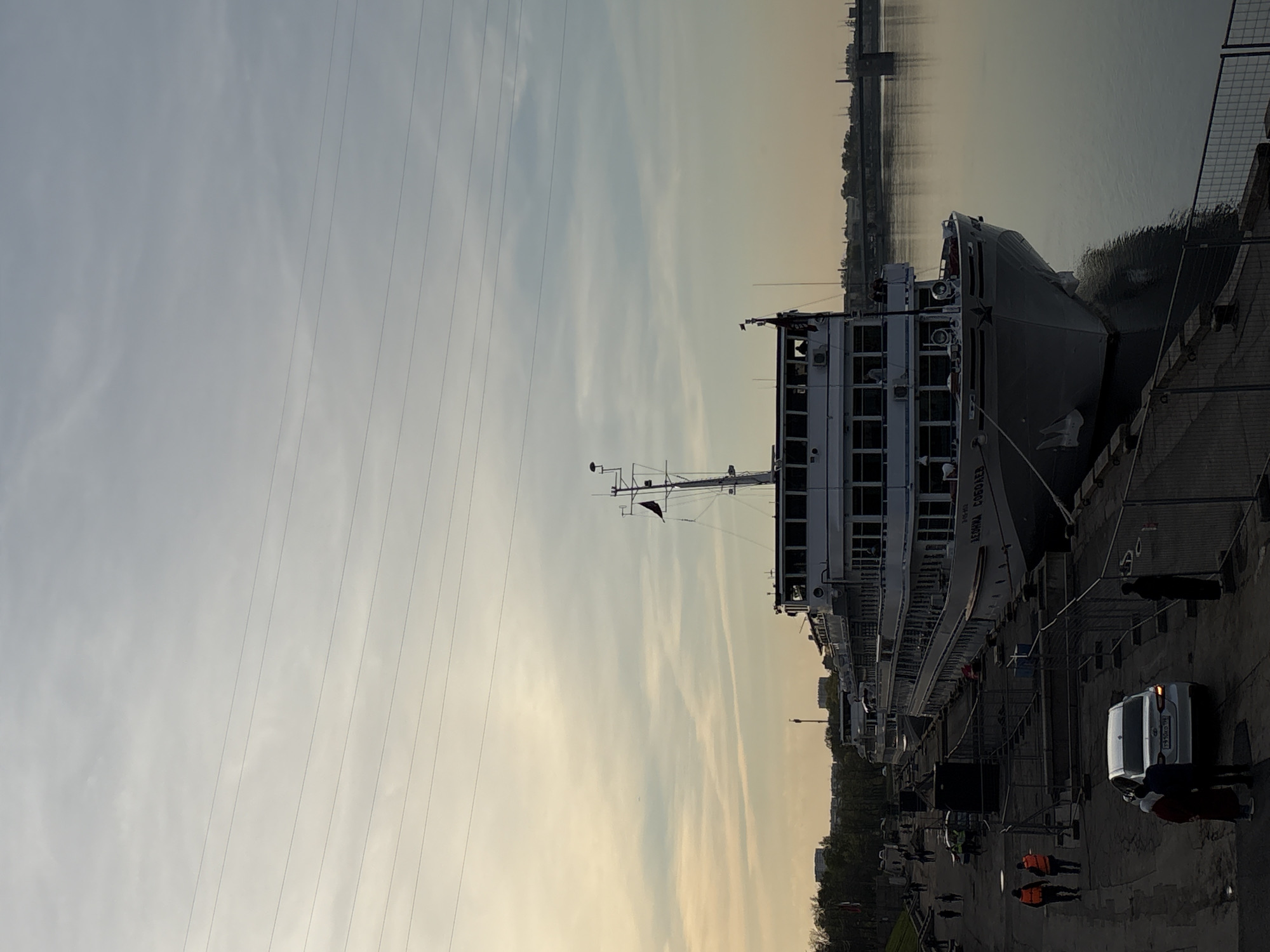 The image depicts a multi-deck river cruise ship, the "Levkova Sobolev," docked along a concrete pier in Saint Petersburg, Russia. A white van is parked near the ship, with people standing around it and walking on the pier. Some people are wearing orange vests. Power lines stretch across the sky in the background, with the cityscape visible in the distance across the water. The sky appears cloudy, with a warm, sunset glow near the horizon. The overall scene suggests a busy waterfront with a docked passenger ship.