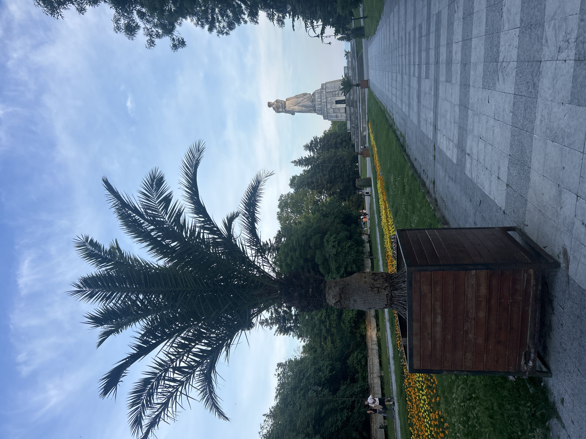 This image depicts an outdoor park or garden scene in the city of Unknown, Unknown, under a partly cloudy blue sky. In the immediate foreground, a mature palm tree with dark green fronds is situated within a rectangular wooden planter with a dark metal frame. To the right of the planter, a paved walkway extends into the distance, characterized by alternating light and dark grey rectangular paving slabs arranged in horizontal stripes. To the left of the walkway and palm tree, a strip of grass is visible, bordered by a long bed of yellow and orange marigold-like flowers.
In the midground, towards the left, two individuals are walking away from the viewer on a parallel path; one appears to be a male in a white t-shirt and dark shorts carrying a white bag, and the other a female in a dark top and light pants. Further back, a large, light-colored stone monument dominates the central view, featuring a standing figure atop a multi-tiered, angular base with a small, dark doorway at its front. Two smaller palm trees are visible on either side of the monument's base. The area surrounding the monument is characterized by green lawns and numerous mature trees, including conifers and deciduous species, forming a dense green backdrop. Two black lampposts with curved arms are visible to the right of the paved walkway, extending towards the monument. The overall lighting suggests daylight, possibly late afternoon, with soft shadows.