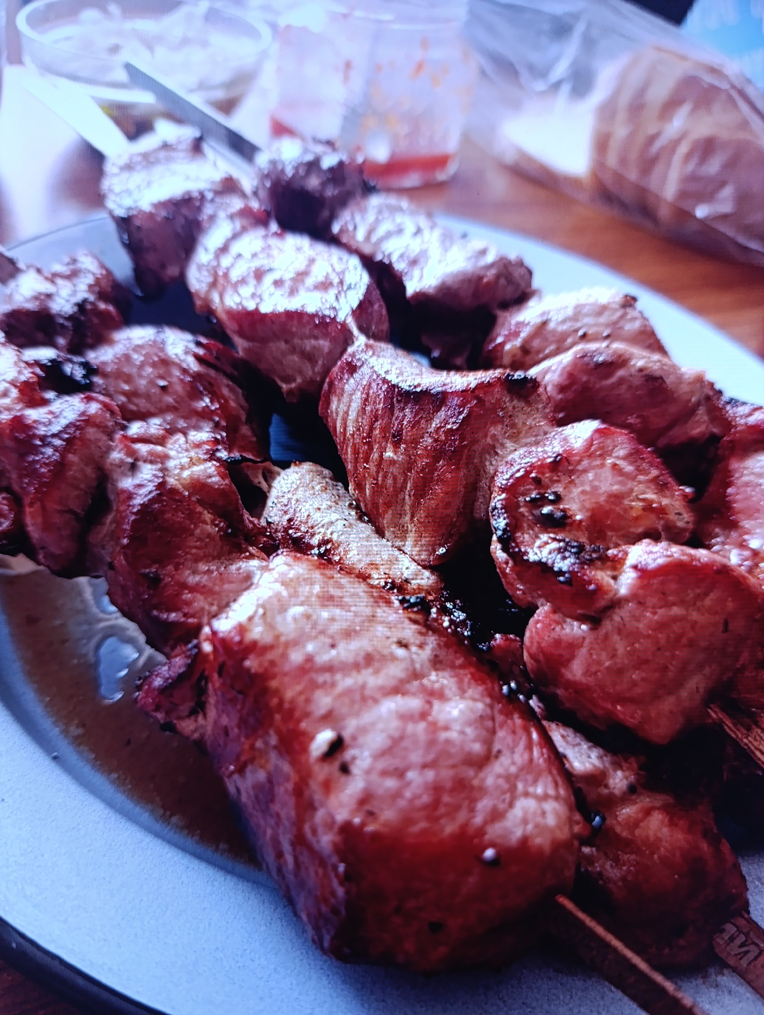 A close-up shot depicts several skewers of cooked meat arranged on a light blue plate. The meat appears to be well-done with some charring. The plate sits on a wooden surface. In the background, there are some blurry containers and what seems to be bread in a plastic bag. There is no evidence of Зипуново, Russia. The city name appears irrelevant to the image content.