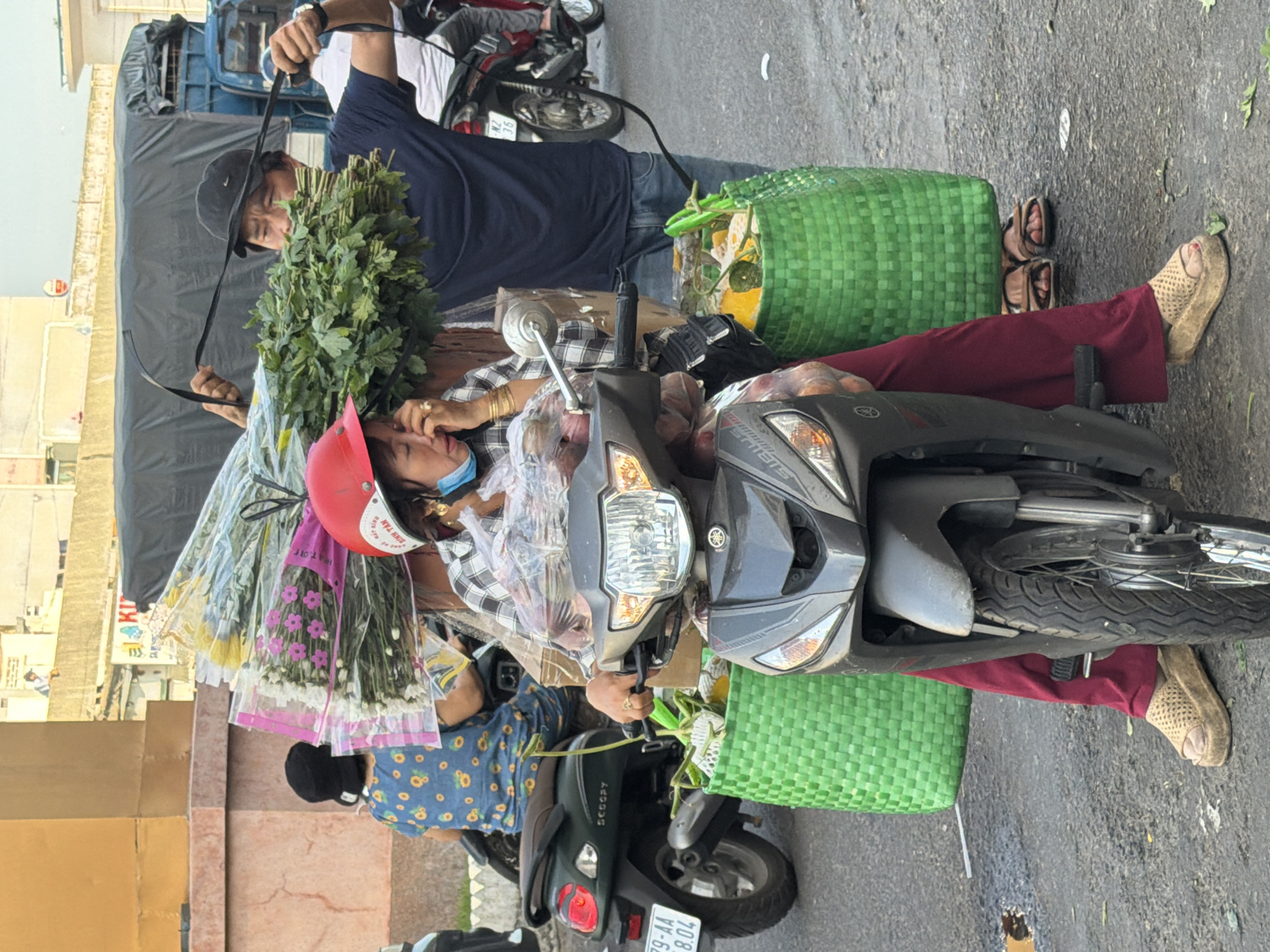 The image shows a street scene in Nha Trang, Vietnam, where two people are transporting a large quantity of flowers and produce on a motorcycle. In the foreground, a woman wearing a red helmet, a plaid shirt, and red pants sits on a dark-colored motorcycle. She is holding the handlebars with her right hand, wearing gold bracelets, and appears to be wiping her eye with her left hand. A light blue face mask is worn below her nose. The motorcycle has two large green woven baskets attached to its sides, filled with produce and possibly more flowers. Behind her, a man wearing a dark blue t-shirt and a black baseball cap is holding a large bunch of green leafy vegetables, secured with black straps attached to the motorcycle. Another person in a blue and yellow sunflower-patterned shirt is visible on a separate scooter in the background, to the left. The environment appears to be an urban street, with buildings and a large truck visible in the background, suggesting a commercial or transportation hub within the city.