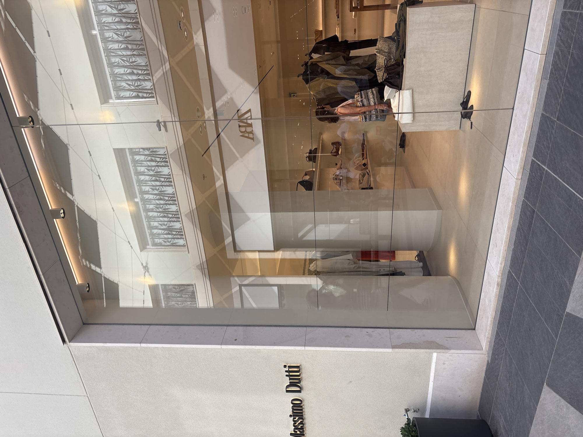 The image shows the storefront of a Massimo Dutti store on the left, with its logo prominently displayed. To the right, through a large glass window, the interior of a Zara store is visible, featuring clothing racks, shelves with handbags, and a woman browsing. The exterior of the buildings suggests an urban environment, possibly a shopping street or mall. The lighting indicates it is daytime, with sunlight reflecting off the glass facade of the Zara store. The ground in front of the stores is paved with dark gray tiles. The overall scene is one of commercial activity and fashion retail. The location is identified as Orihuela, Spain, though this specific context is not visually verifiable from the image alone.
